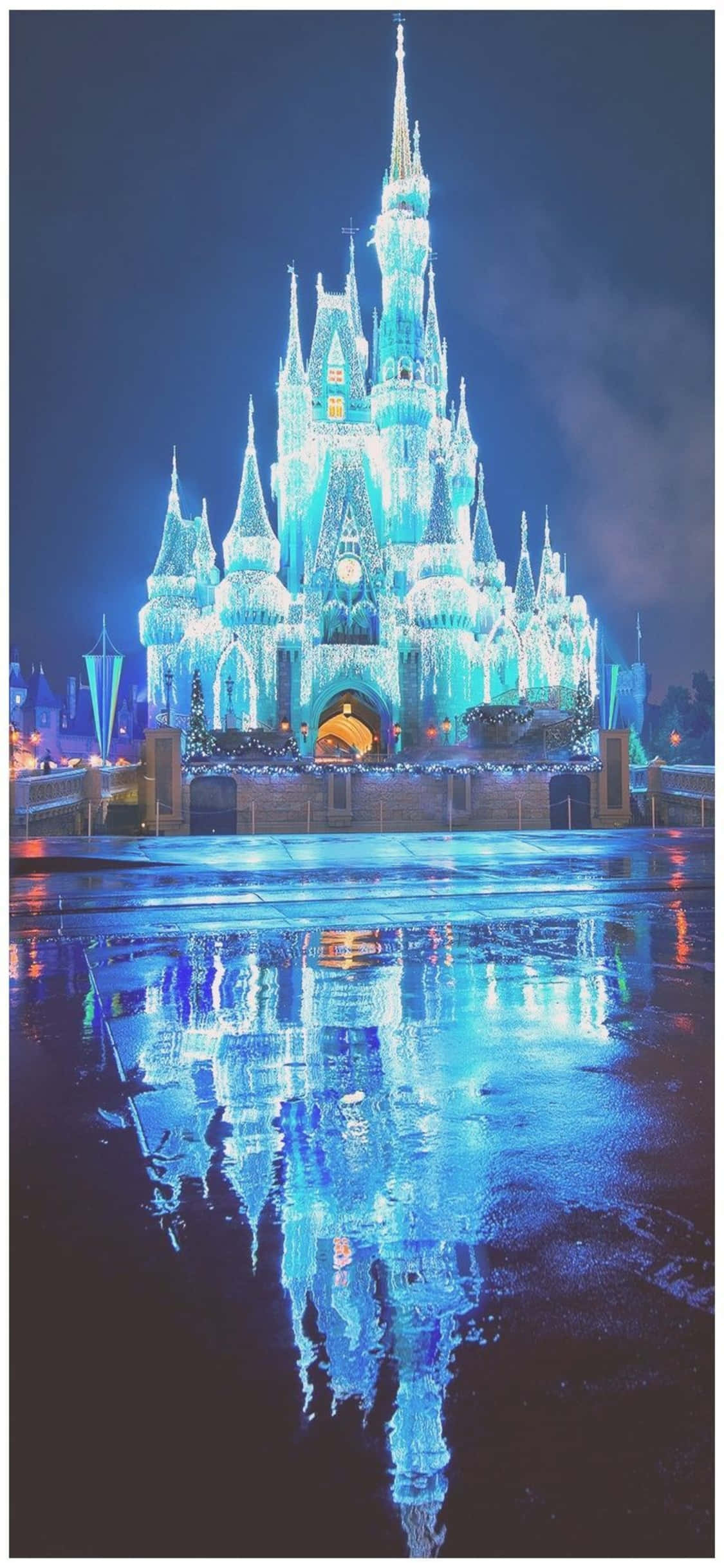 walt disney 桌布 壁紙 — 一座雄偉的城堡在鮮豔的藍色燈光照射下，完美地倒映在黑暗夜空下的濕漉漉路面上。