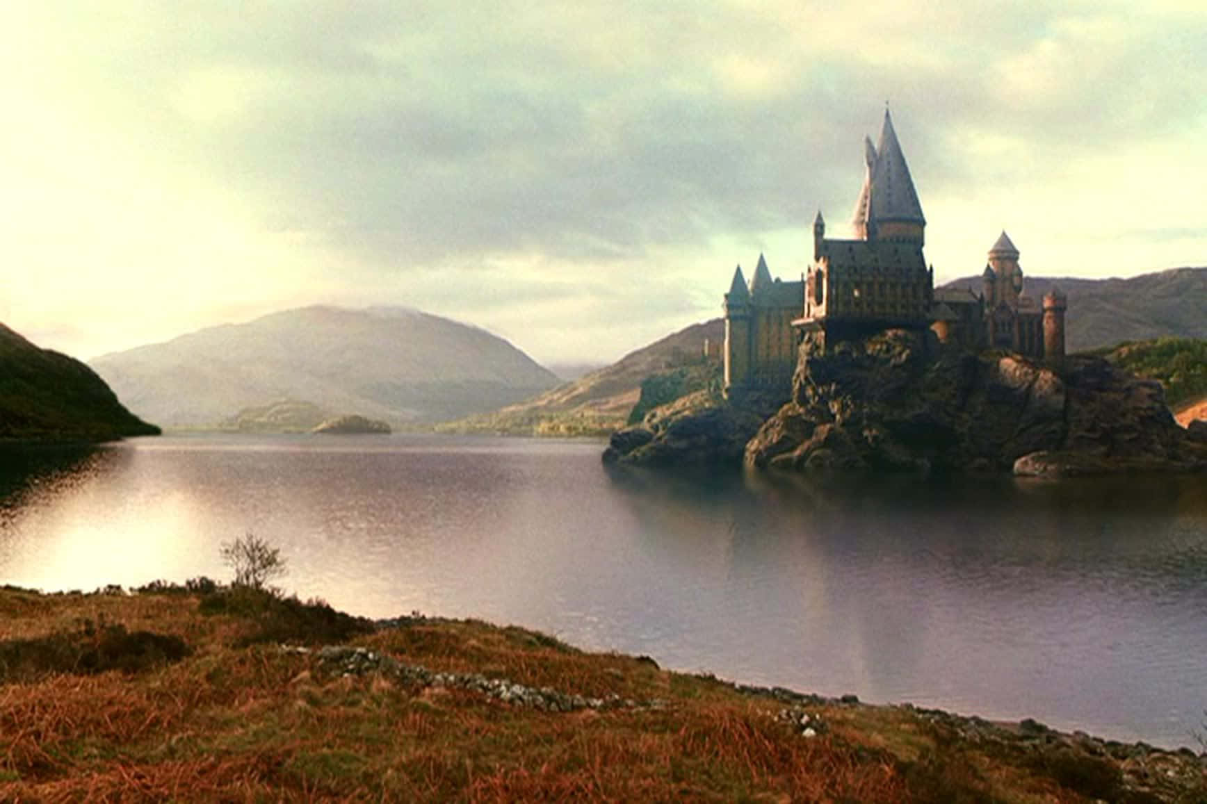 The Hogwarts Lake 桌布 壁紙 — 一座雄偉的城堡坐落在寧靜湖泊中的岩石島上，周圍環繞著起伏的丘陵，天空呈現柔和的金色。
