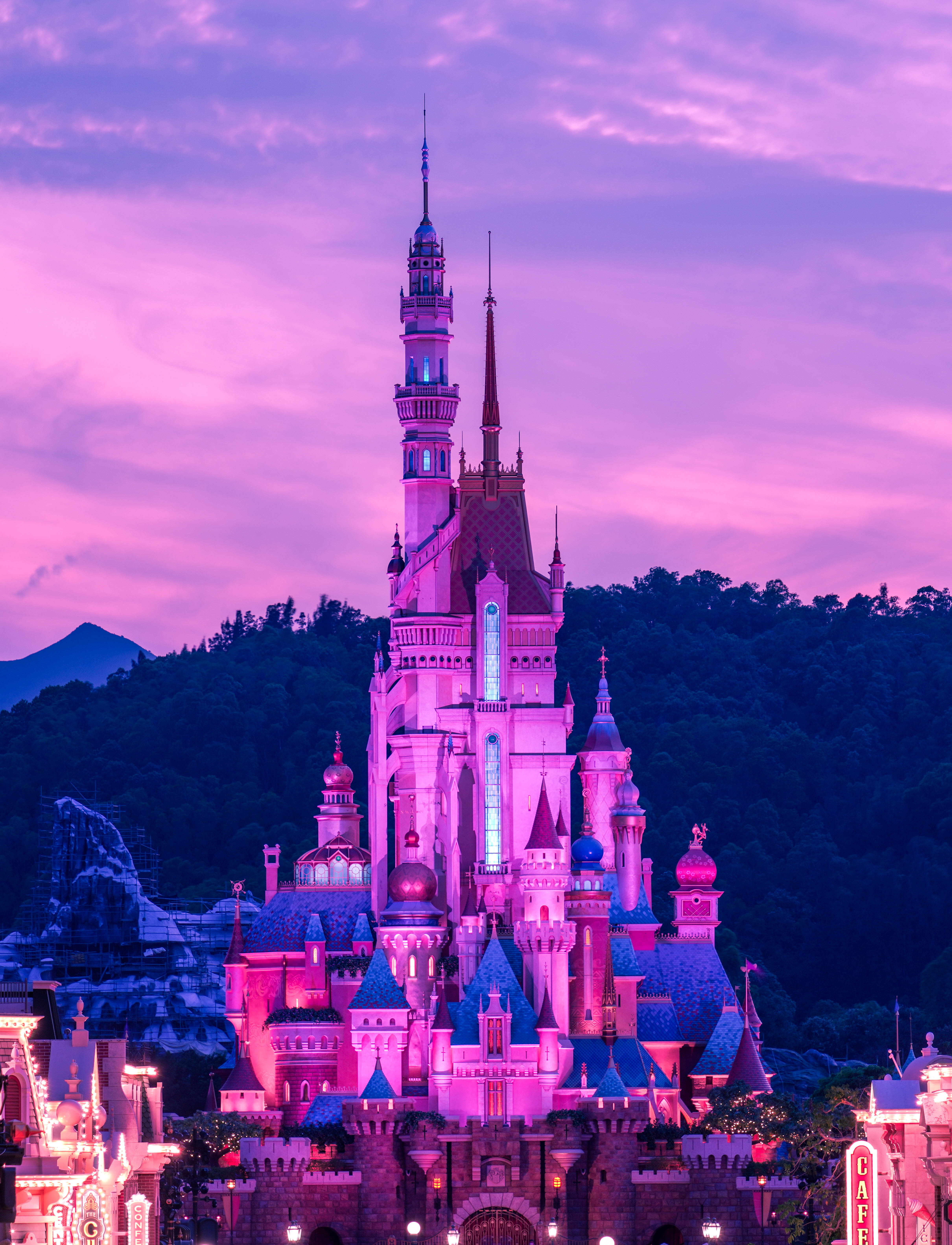 Disneyland Desktop 桌布 壁紙 — 一座雄偉的城堡沐浴在充滿活力的粉紅色和紫色燈光中，背景是暮色天空和遠處的山脈。