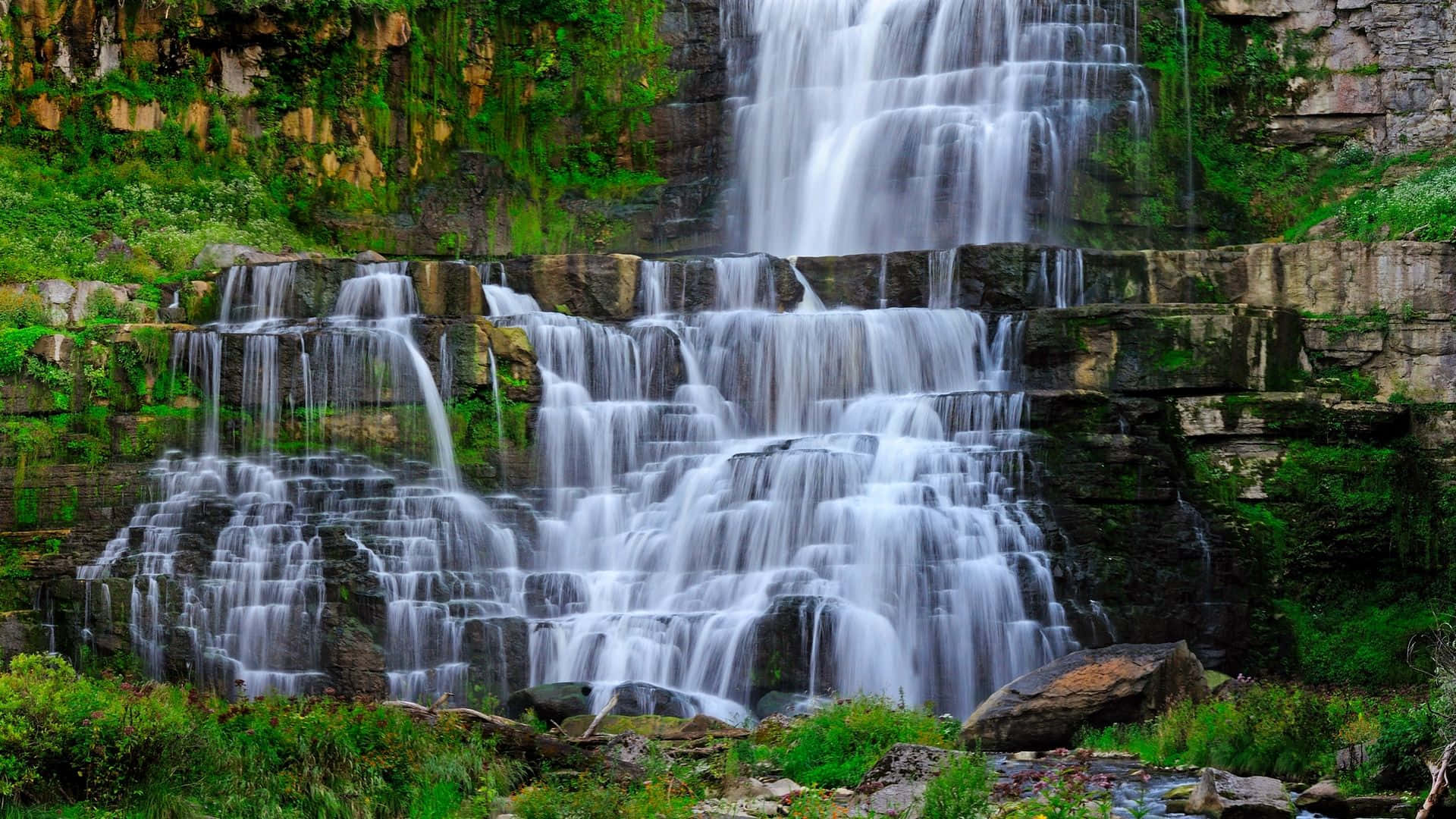 Waterfalls 桌布 壁紙 — 一座雄偉的多層瀑布在明亮的日光下，從長滿苔蘚的岩石上傾瀉而下，周圍環繞著茂密的綠植。