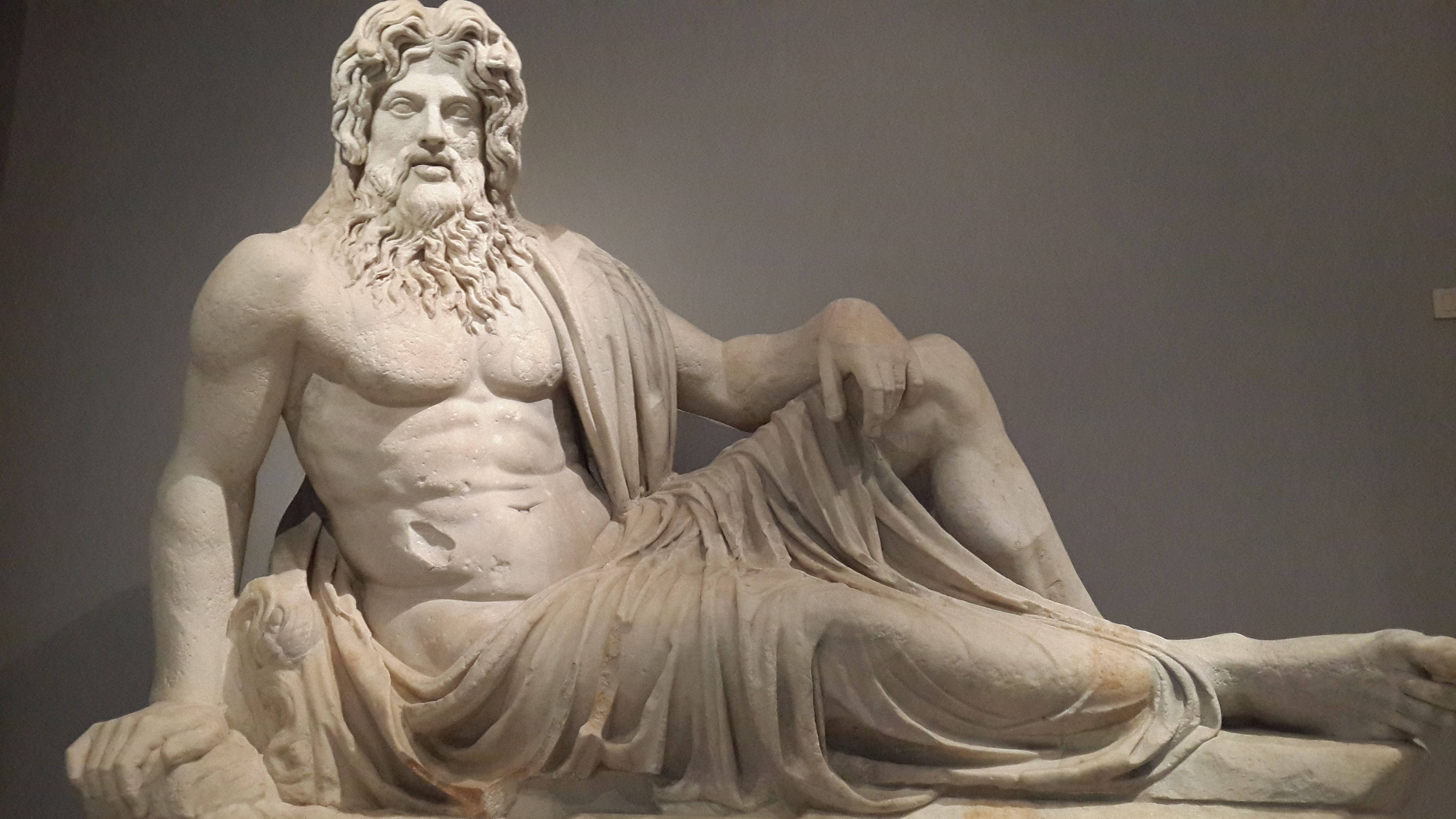 Statue Aesthetic 桌布 壁紙 — 一座雄偉的大理石鬍鬚神雕像，優雅地披著飄逸長袍，在博物館環境中斜倚。