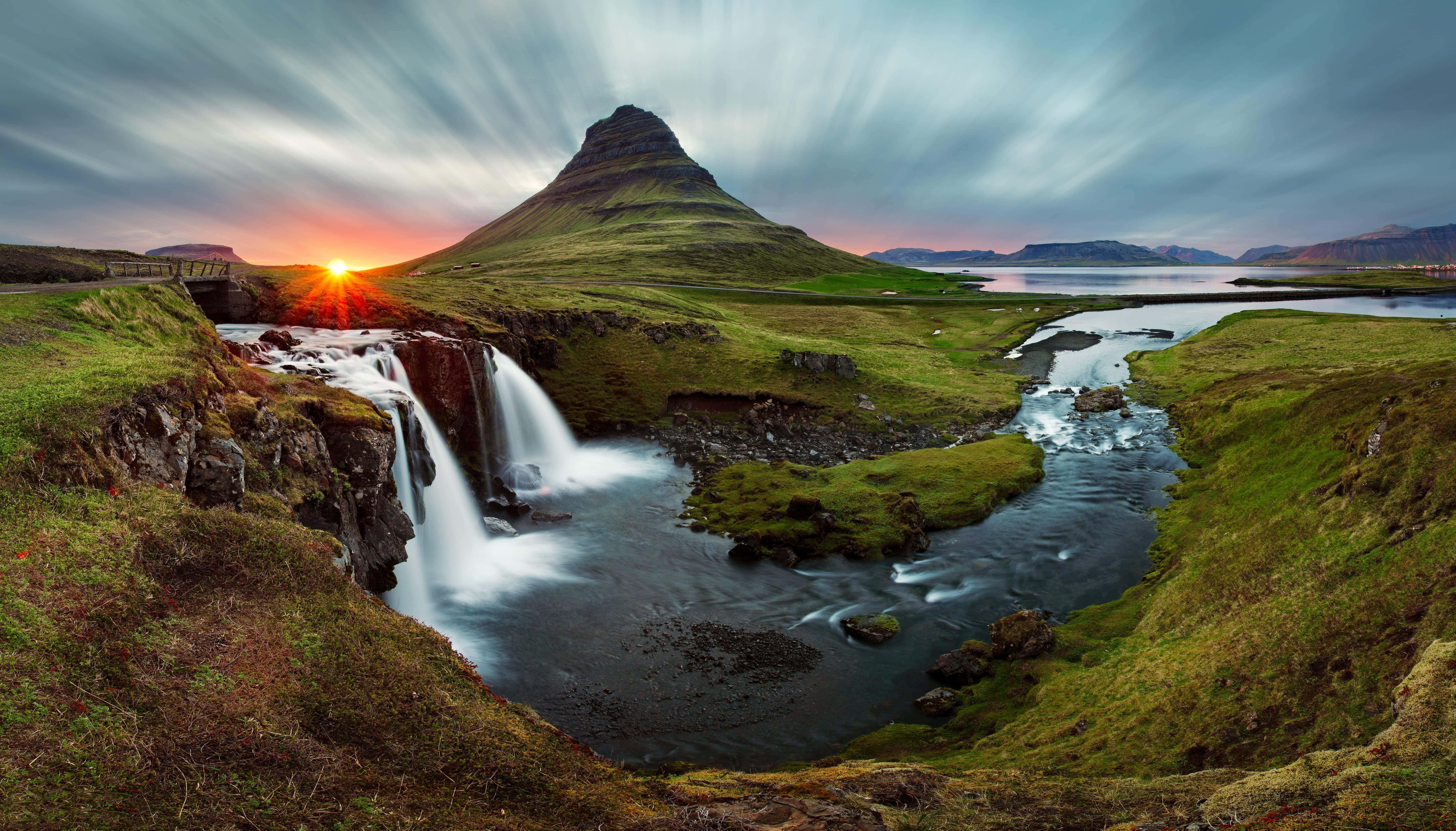 Iceland Desktop 桌布 壁紙 — 一座雄偉的山峰在瀑布後面升起，夕陽西下，將溫暖的光芒灑在綠色的山谷和河流上。