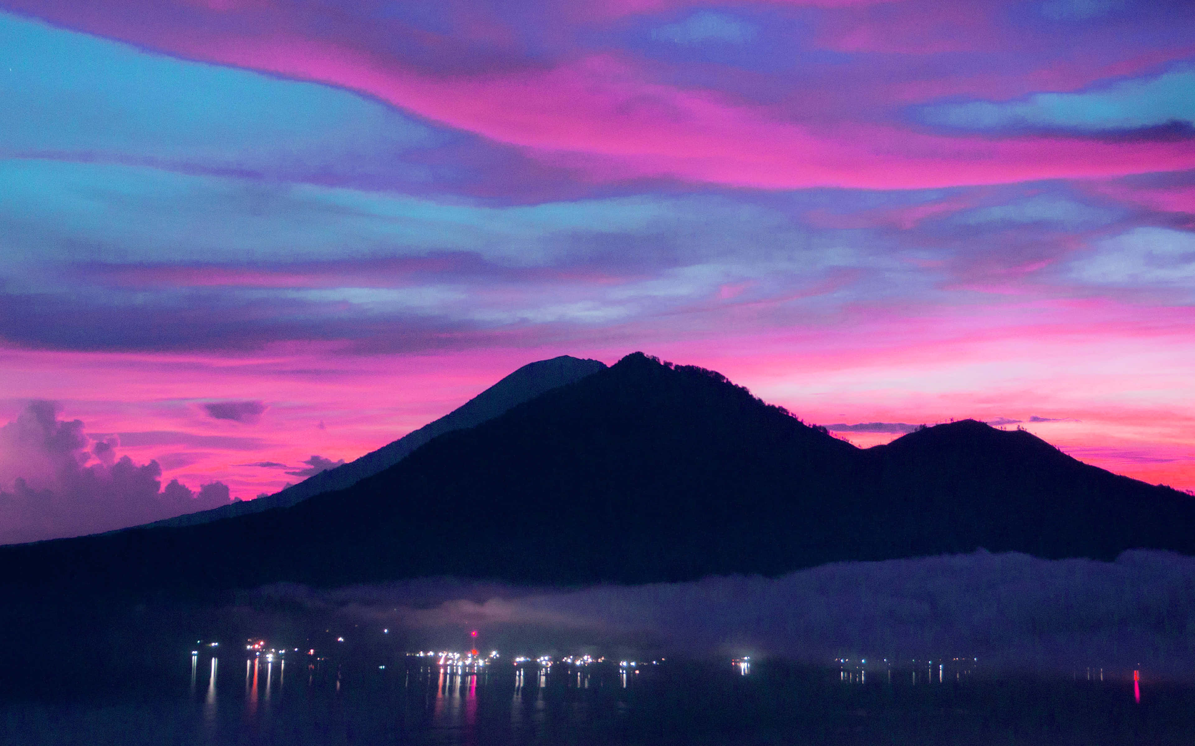 Mountains Sunset 桌布 壁紙 — 一座雄偉的山脈剪影矗立在充滿活力的粉紅色和藍色暮色天空中，下方水面上有遠處燈光的朦朧倒影。