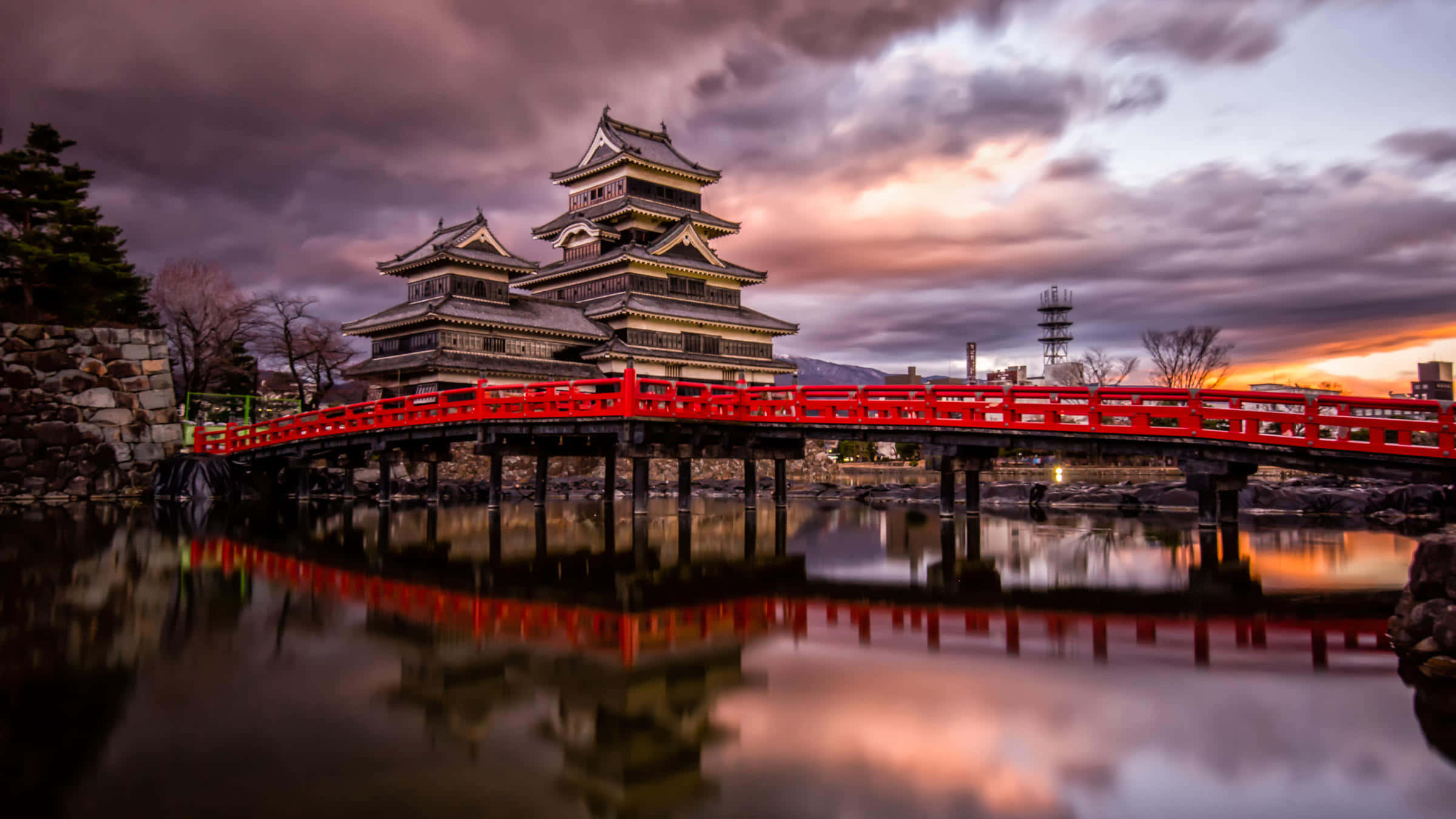 城堡動畫 桌布 壁紙 — 一座雄偉的日本城堡，擁有層疊屋頂，矗立在一座鮮紅色的橋樑後方，橋樑倒映在平靜的水面上，背景是戲劇性的黃昏天空。