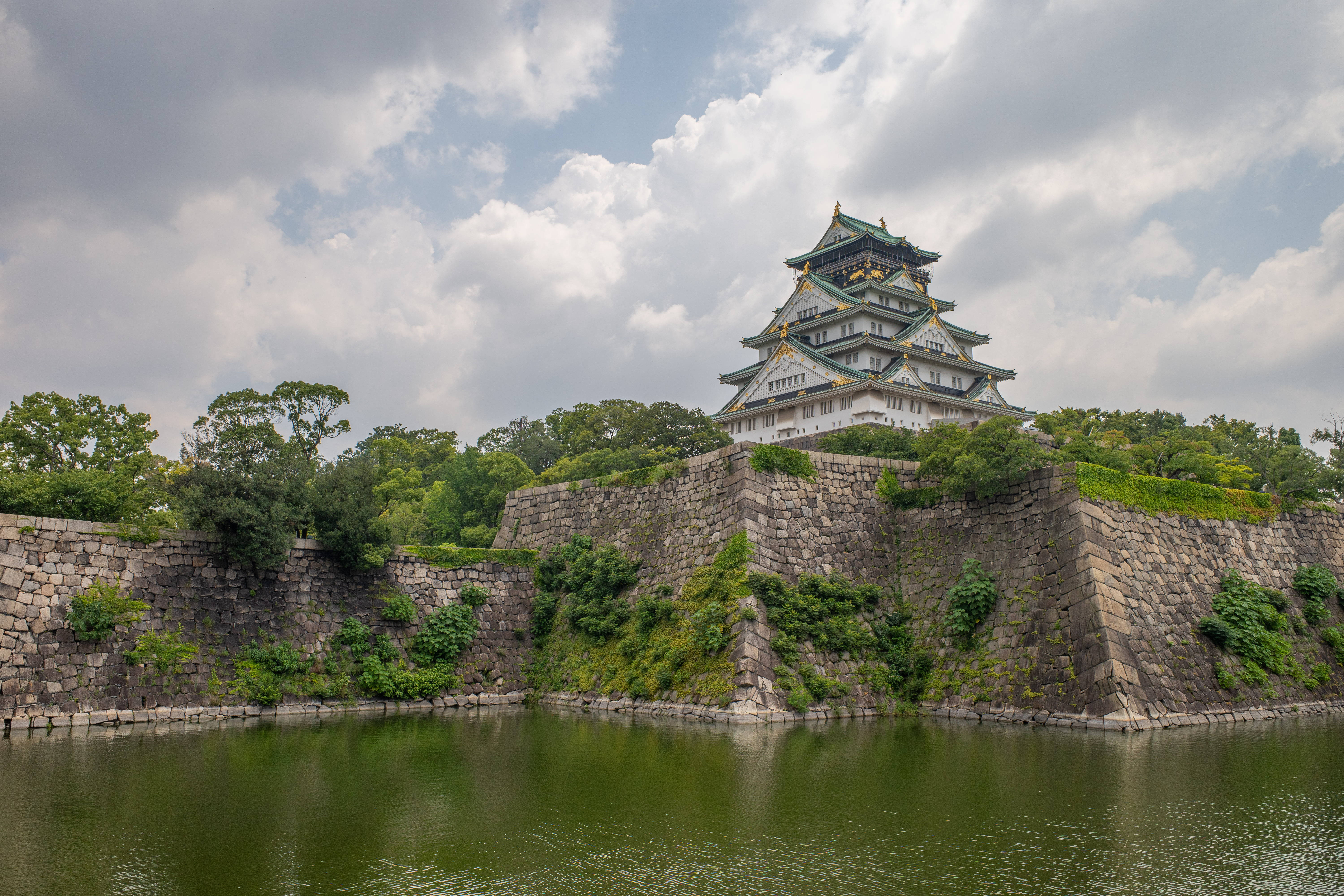 Osaka Castle 桌布 壁紙 — 一座雄偉的日本城堡，擁有綠色屋頂和白色牆壁，矗立在石牆上，周圍環繞著平靜的護城河，天空多雲。