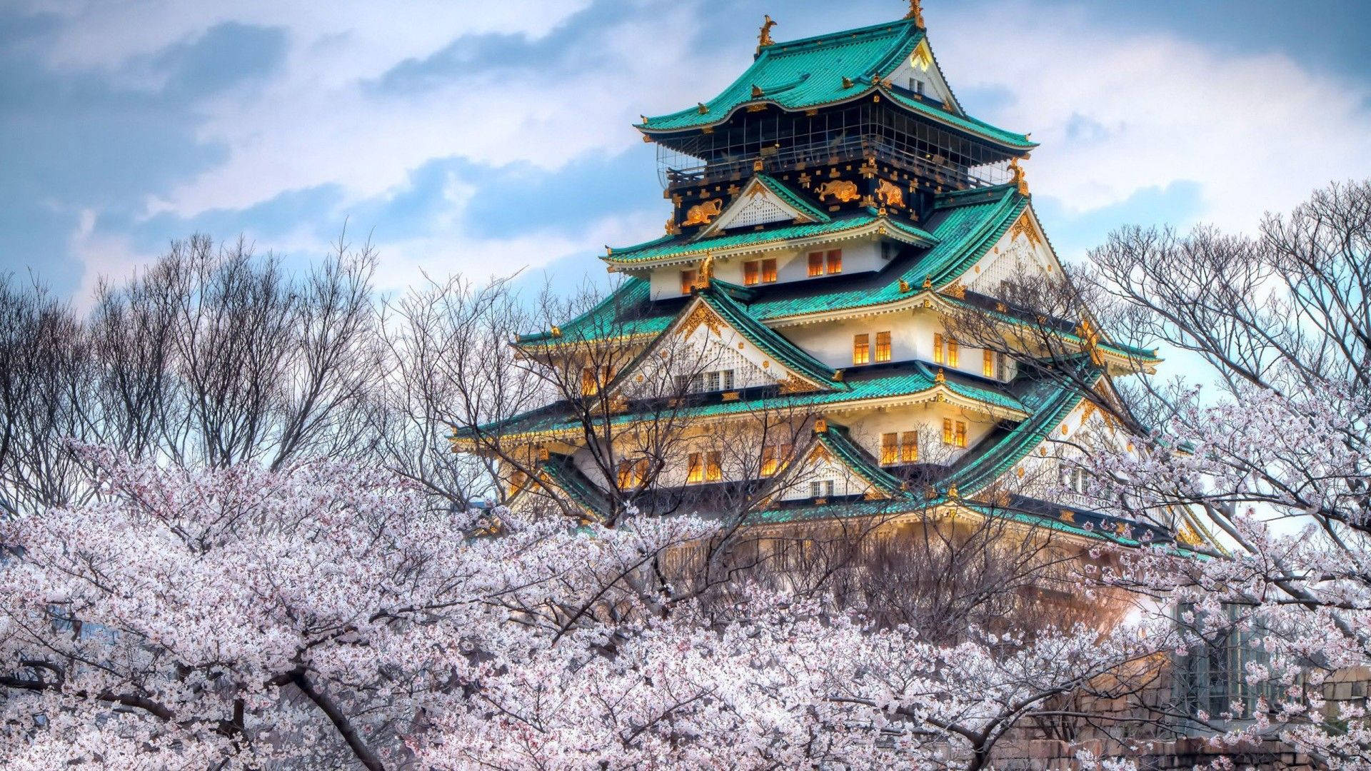 Nagoya 桌布 壁紙 — 一座雄偉的日本城堡，擁有綠色屋頂和發光的窗戶，在陰雲密布的天空下，從盛開的粉紅色櫻花後方升起。
