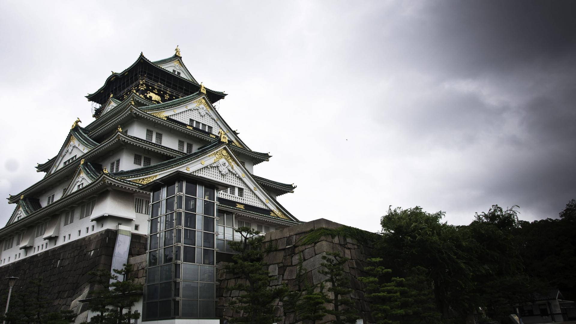 Osaka Castle 桌布 壁紙 — 一座雄偉的日本城堡，白色牆壁和金色點綴，矗立在戲劇性的灰色天空下，周圍環繞著石牆和綠樹。