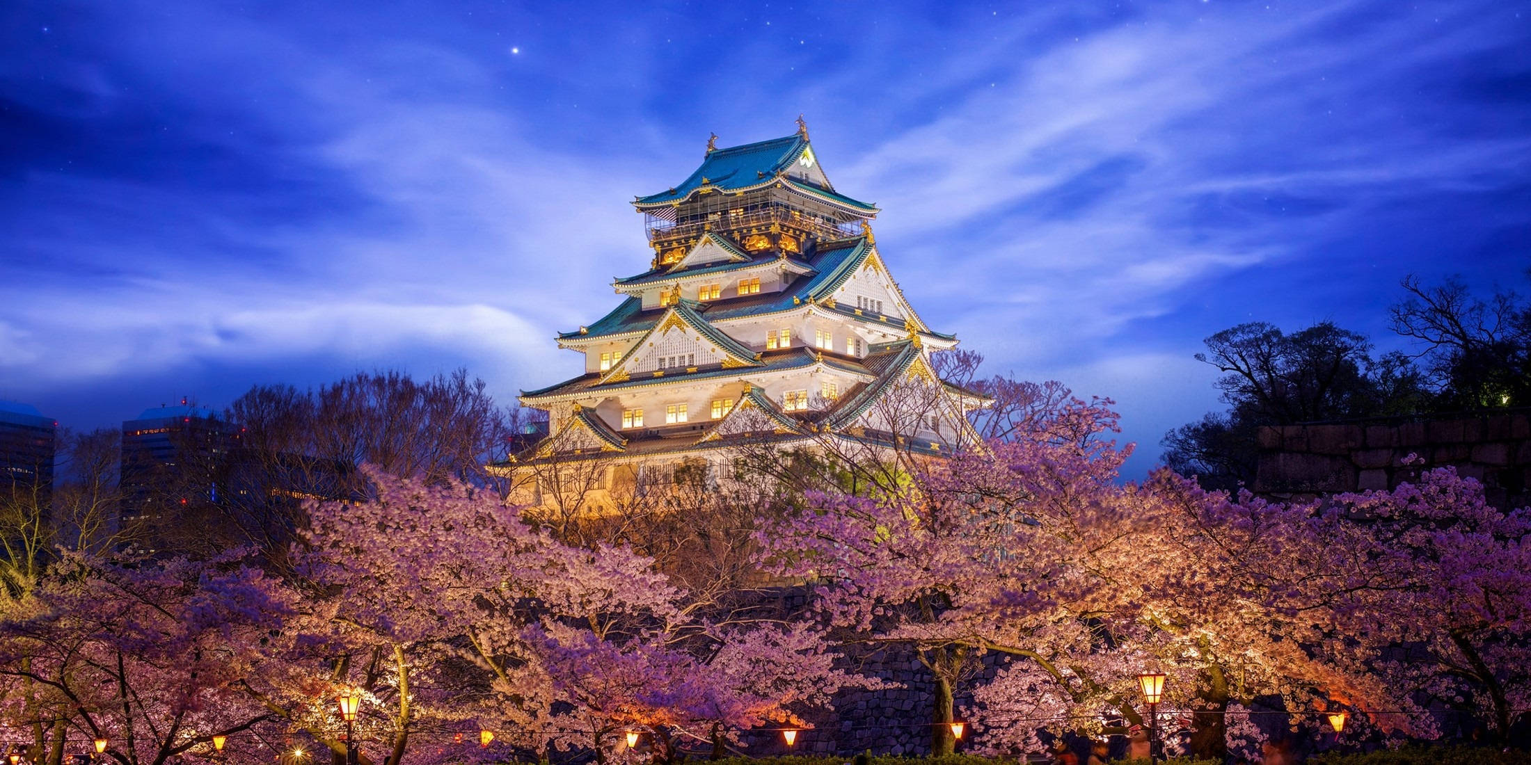 Osaka Castle 桌布 壁紙 — 一座雄偉的日本城堡在黃昏時分被照亮，在繁星點點的藍天下，以盛開的粉紅色櫻花為框。