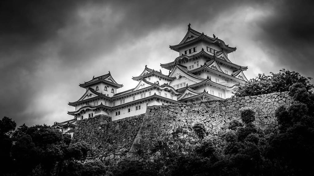 Himeji Castle 桌布 壁紙 — 一座雄偉的日本城堡坐落在石山上，單色調的戲劇性多雲天空下。