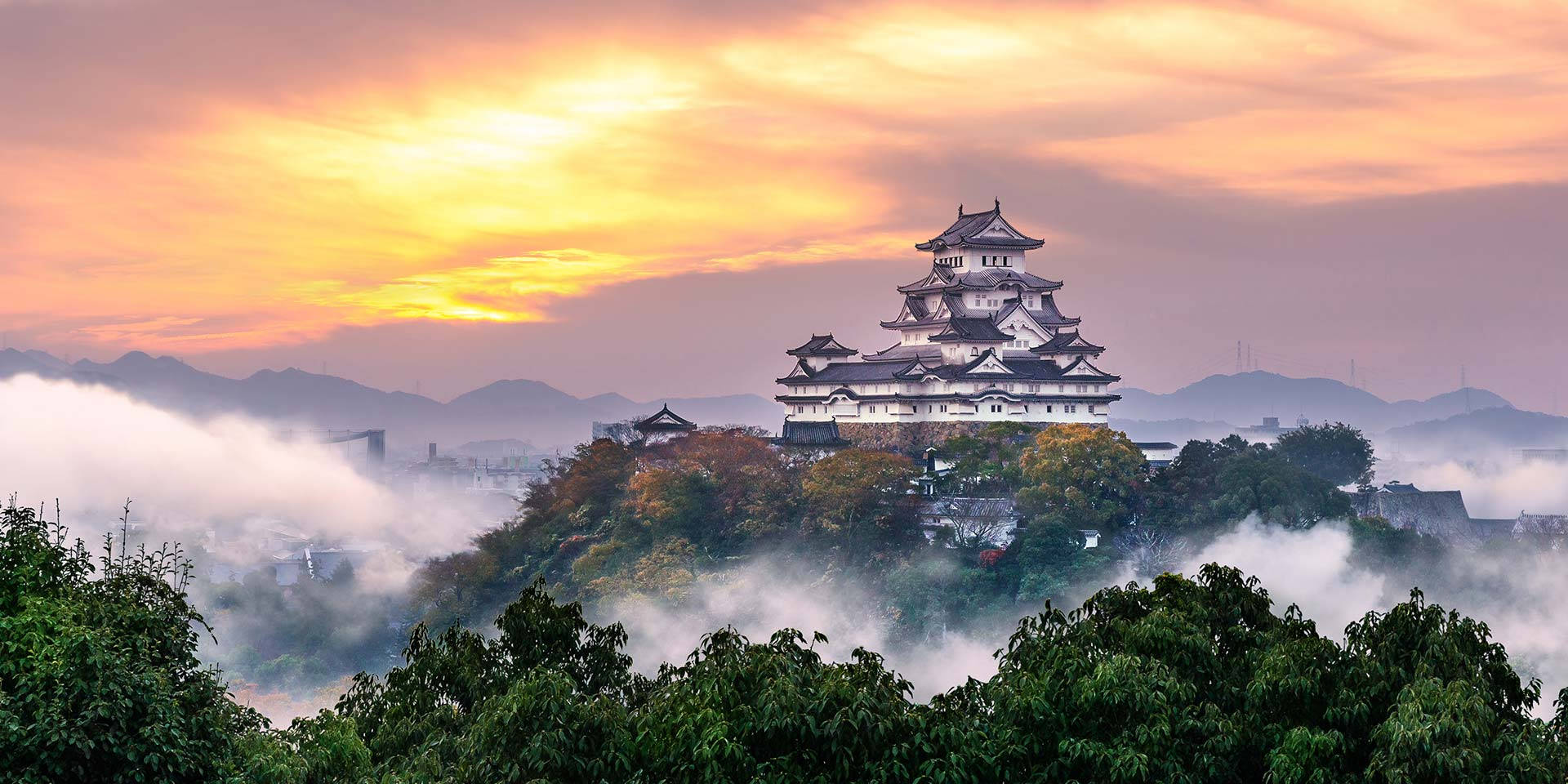 Himeji Castle 桌布 壁紙 — 一座雄偉的日本城堡坐落在霧氣瀰漫的山丘上，日出時分，金色雲彩和秋樹環繞其下。