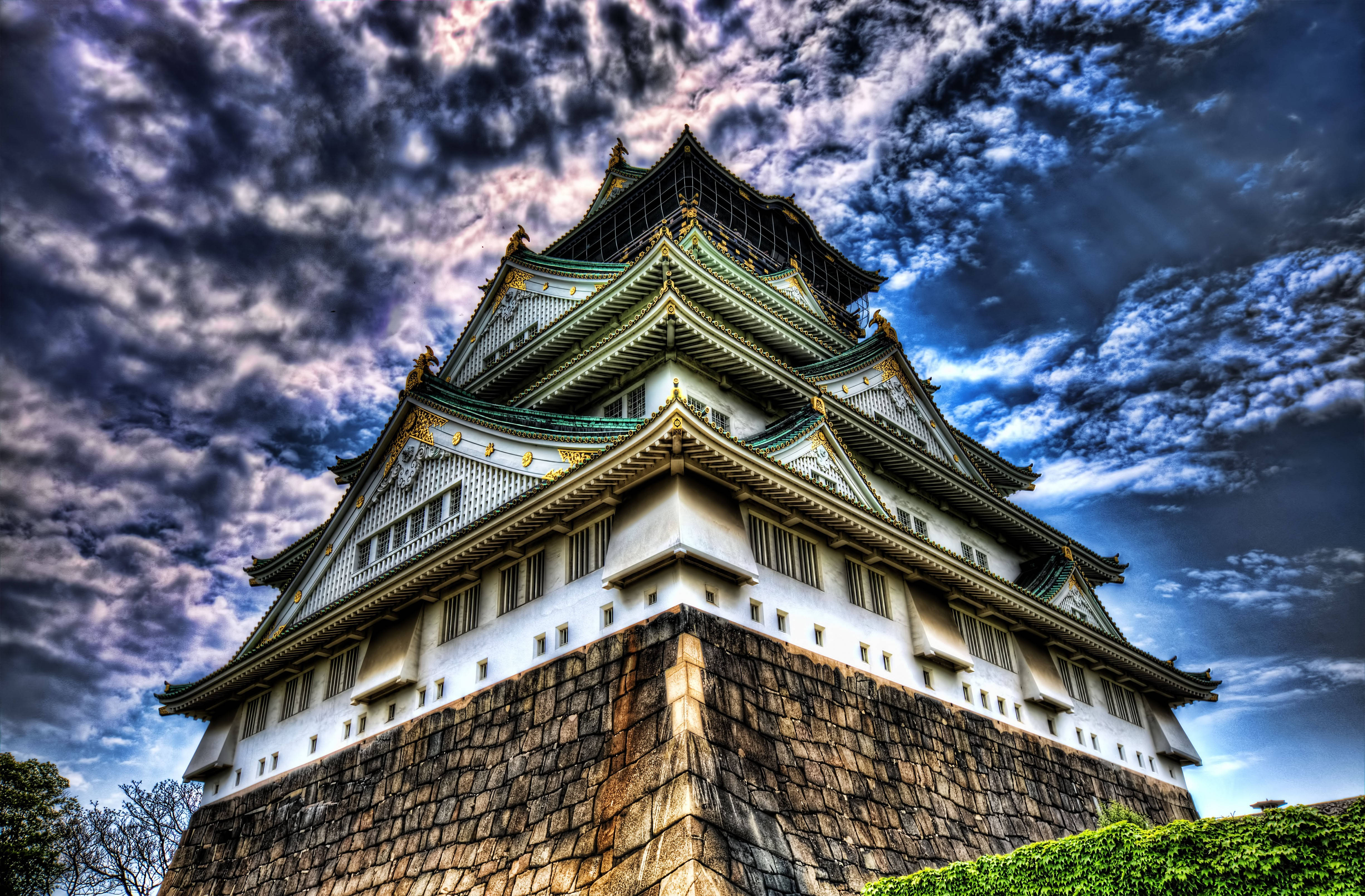 Osaka Castle 桌布 壁紙 — 一座雄偉的日本城堡塔樓，擁有綠色瓦片屋頂和白色牆壁，在戲劇性的多雲天空下巍然聳立。