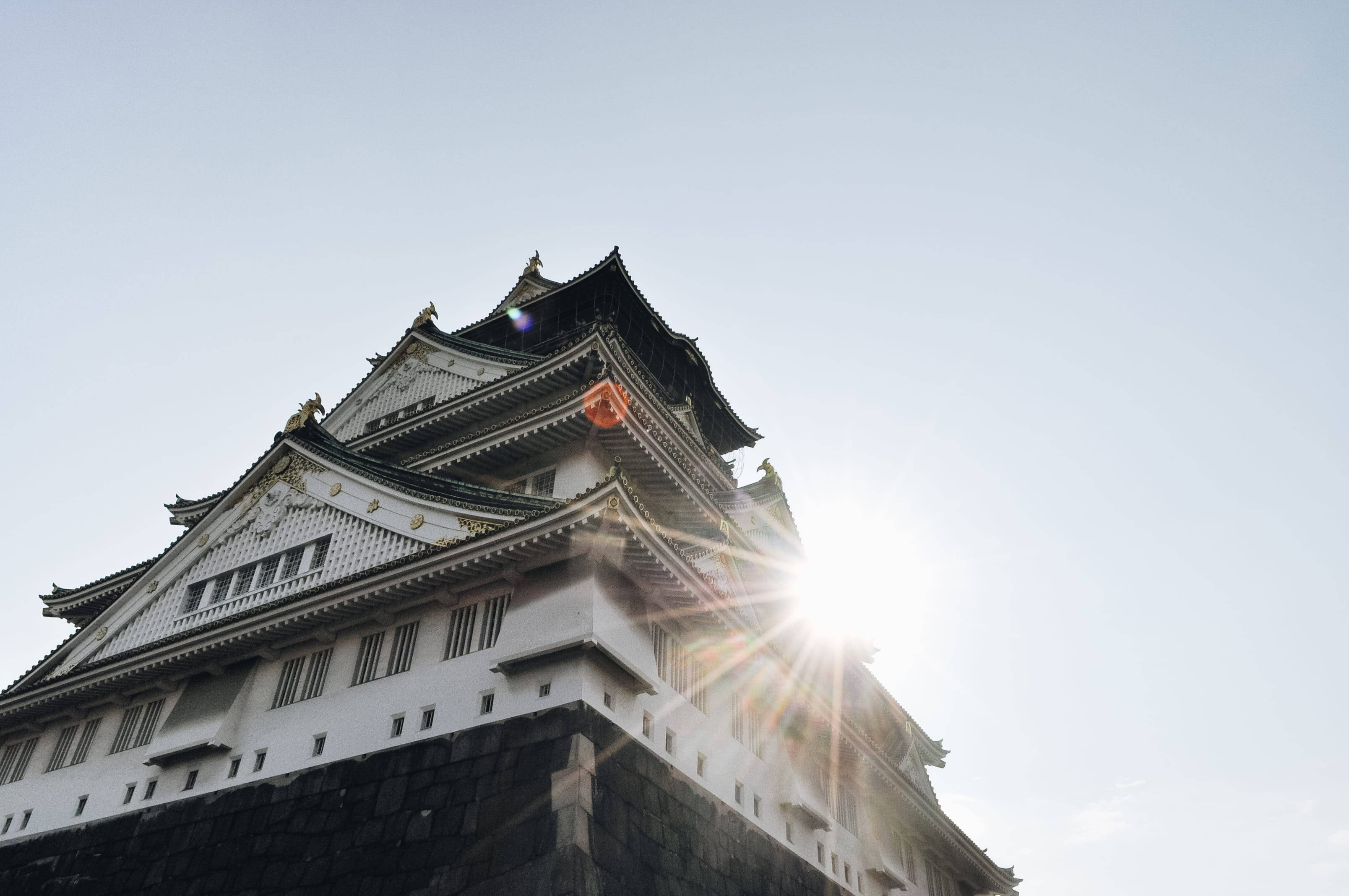 Osaka Castle 桌布 壁紙 — 一座雄偉的日本城堡塔樓在晴朗的天空下聳立，太陽從其屋頂線穿透而出，產生燦爛的鏡頭光暈。