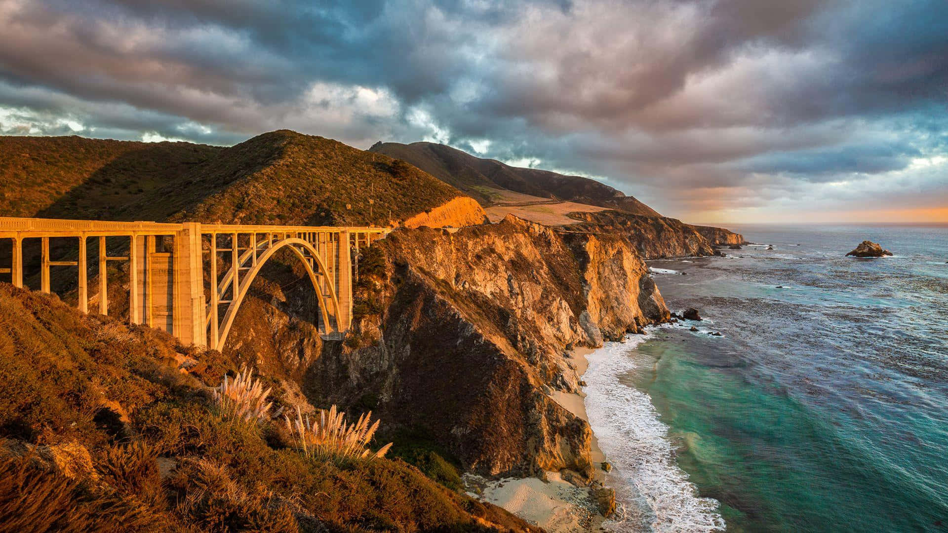 Big Sur 桌布 壁紙 — 一座雄偉的海岸橋樑在崎嶇的懸崖上拱起，金色的夕陽光芒以溫暖的色調沐浴著場景，映襯著戲劇性的多雲天空。