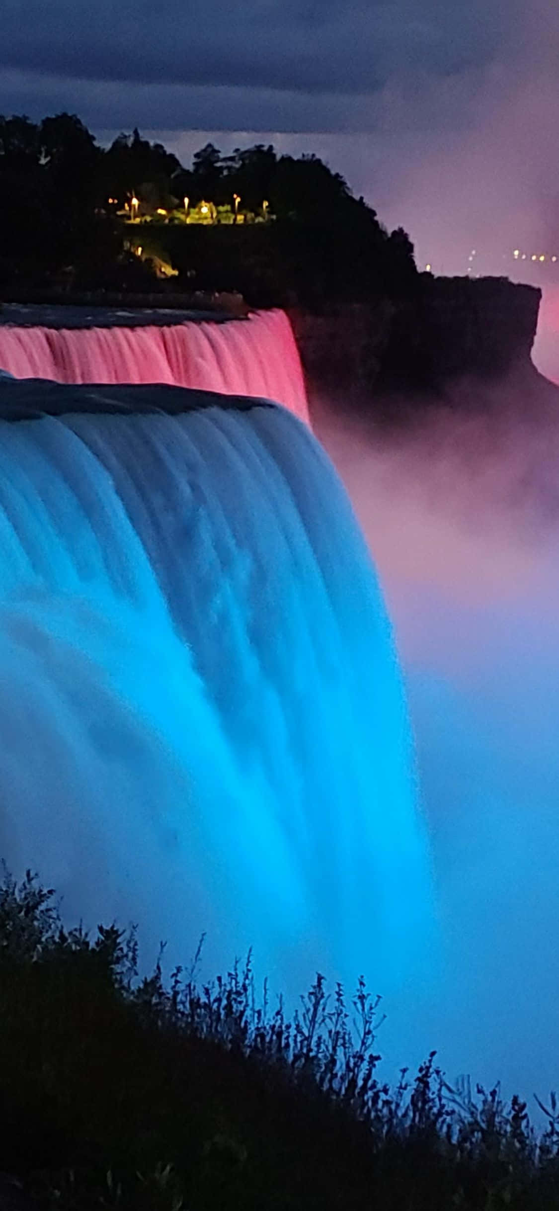 Niagara Falls 桌布 壁紙 — 一座雄偉的瀑布在夜間被鮮豔的藍色和粉紅色燈光照亮，壯觀地傾瀉而下，薄霧在其底部旋轉。