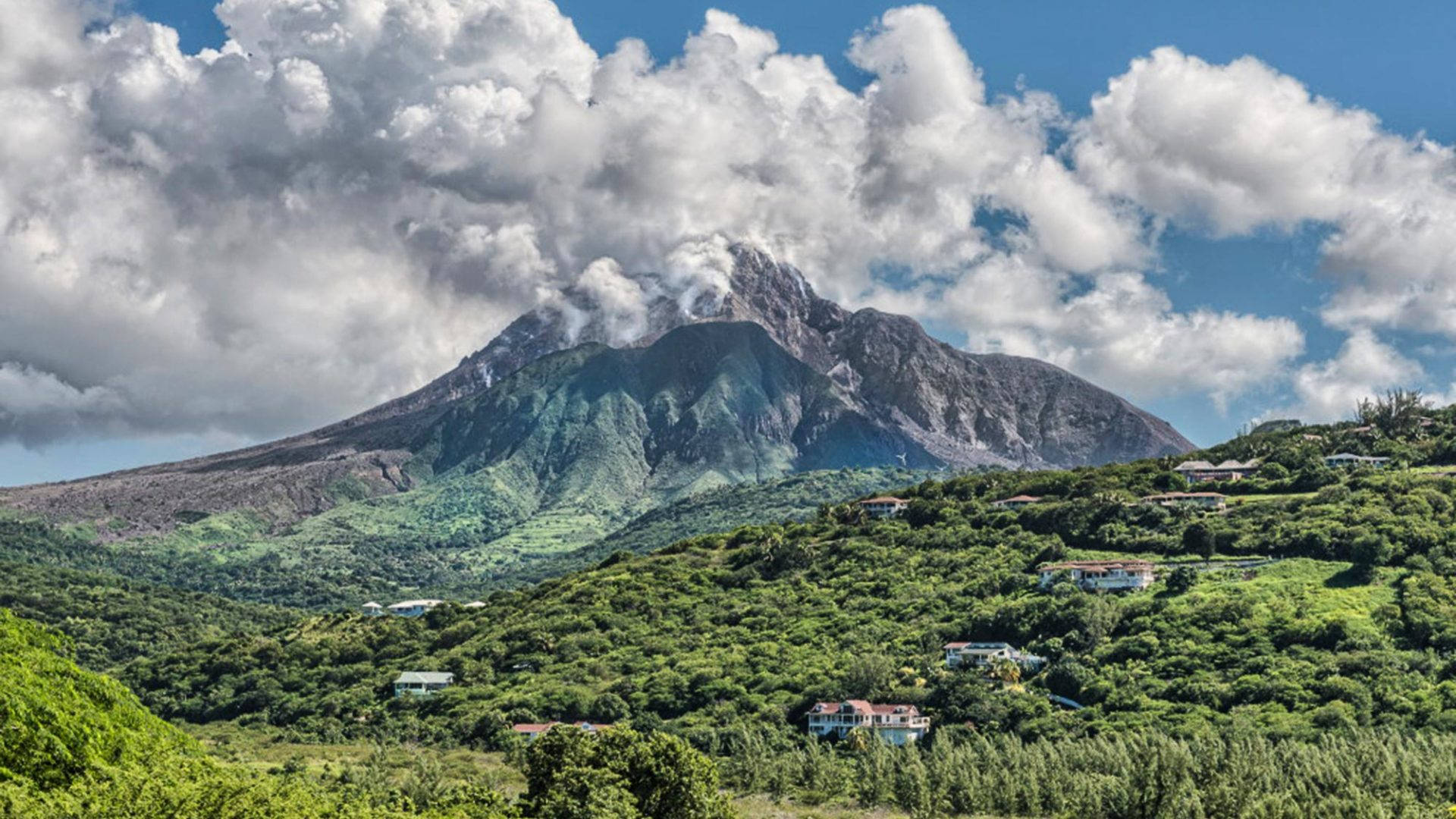 森林 桌布 壁紙 — 一座雄偉的火山冒出一縷煙霧，聳立在鬱鬱蔥蔥的綠色山丘上，山丘上點綴著色彩繽紛的房屋，天空部分多雲，呈現藍色。