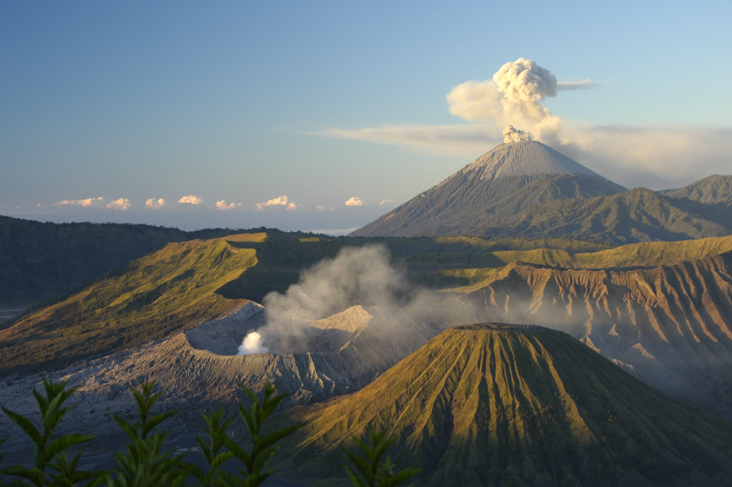 印尼 桌布 壁紙 — 一座雄偉的火山噴發，煙霧升騰，映襯著清澈的藍天，金色的陽光灑滿崎嶇的地貌。