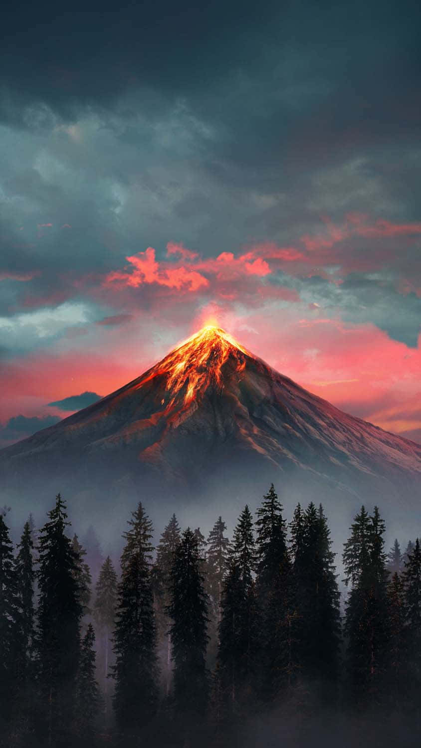 山景 桌布 壁紙 — 一座雄偉的火山噴發，發出熾熱的光芒，映襯著暮色天空，粉紅色和灰色的雲彩劃過，下方是霧氣瀰漫的松樹林。