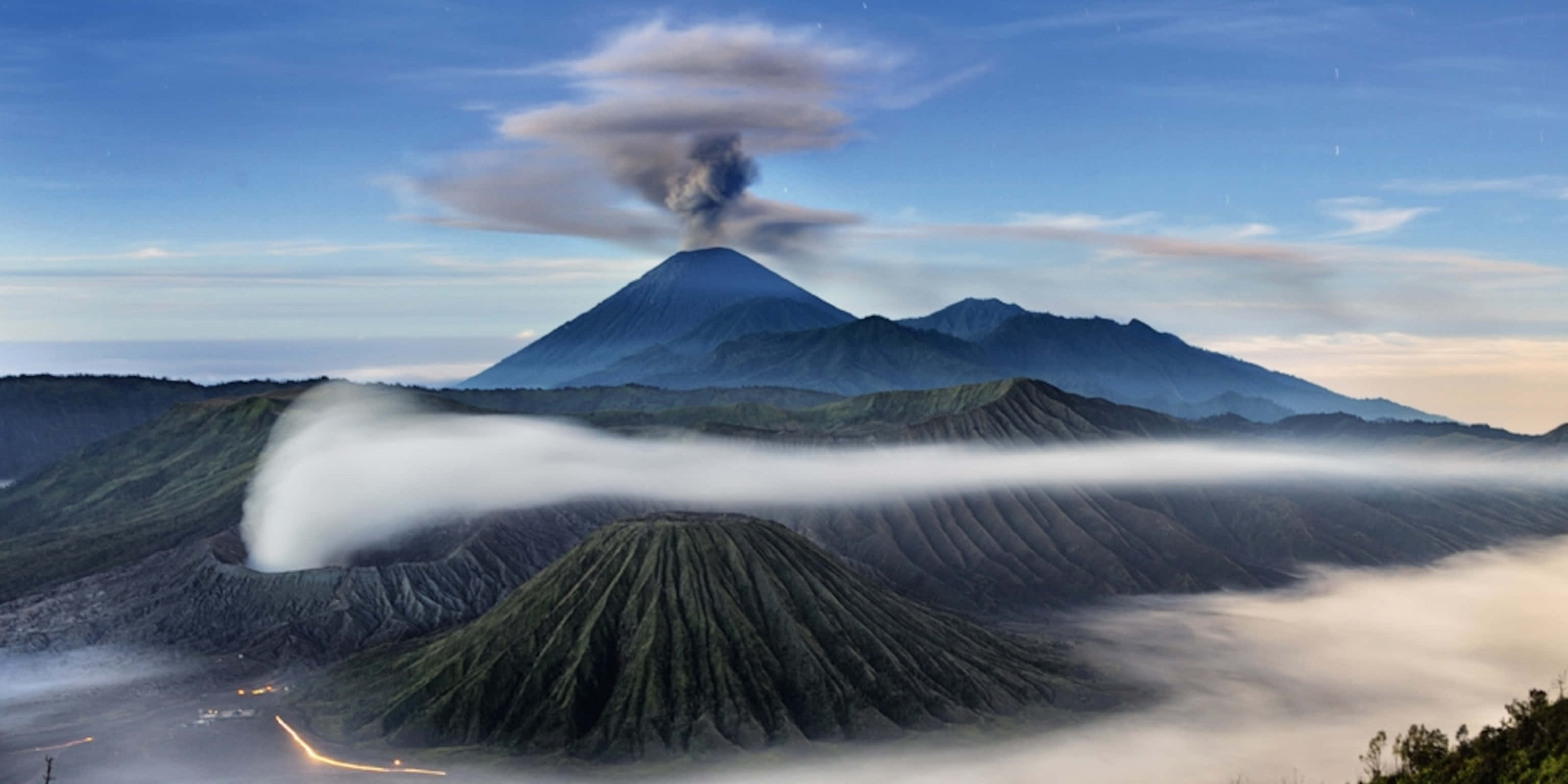 山景 桌布 壁紙 — 一座雄偉的火山噴發著黑煙，同時厚厚的白雲流過下方霧氣瀰漫的山谷。