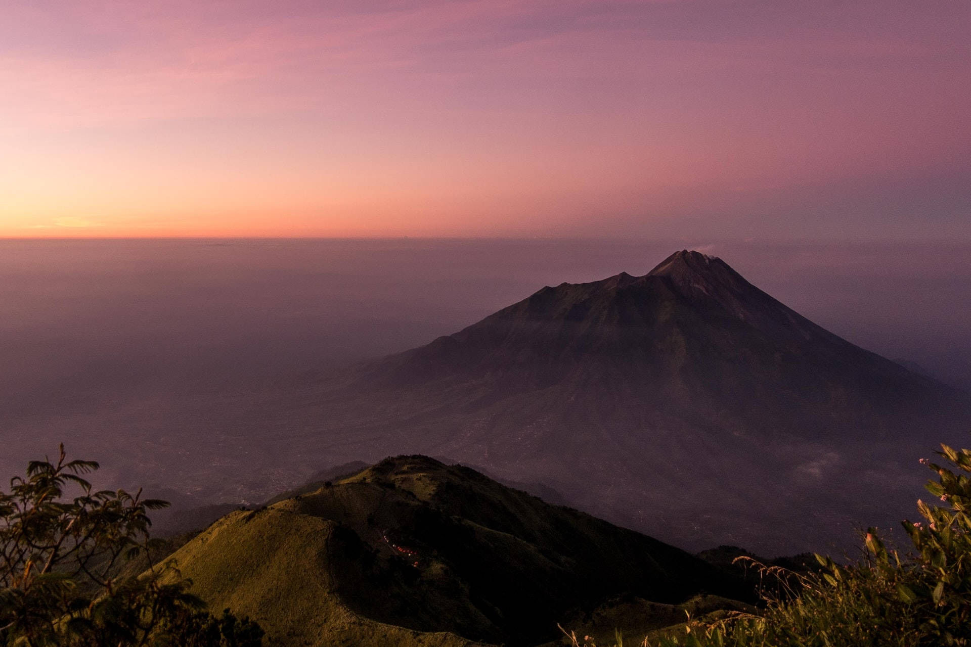 Mountain Landscape 桌布 壁紙 — 一座雄偉的火山在柔和的粉紅色和橙色黃昏天空下聳立，下方是霧氣瀰漫的山谷。