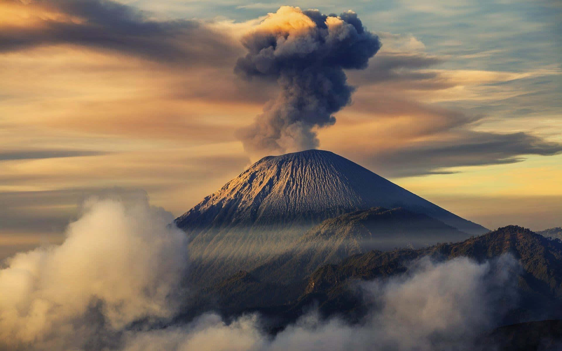 印尼 桌布 壁紙 — 一座雄偉的火山在金色日出天空的映襯下噴發著黑煙，周圍環繞著霧氣繚繞的雲朵和崎嶇的山峰。