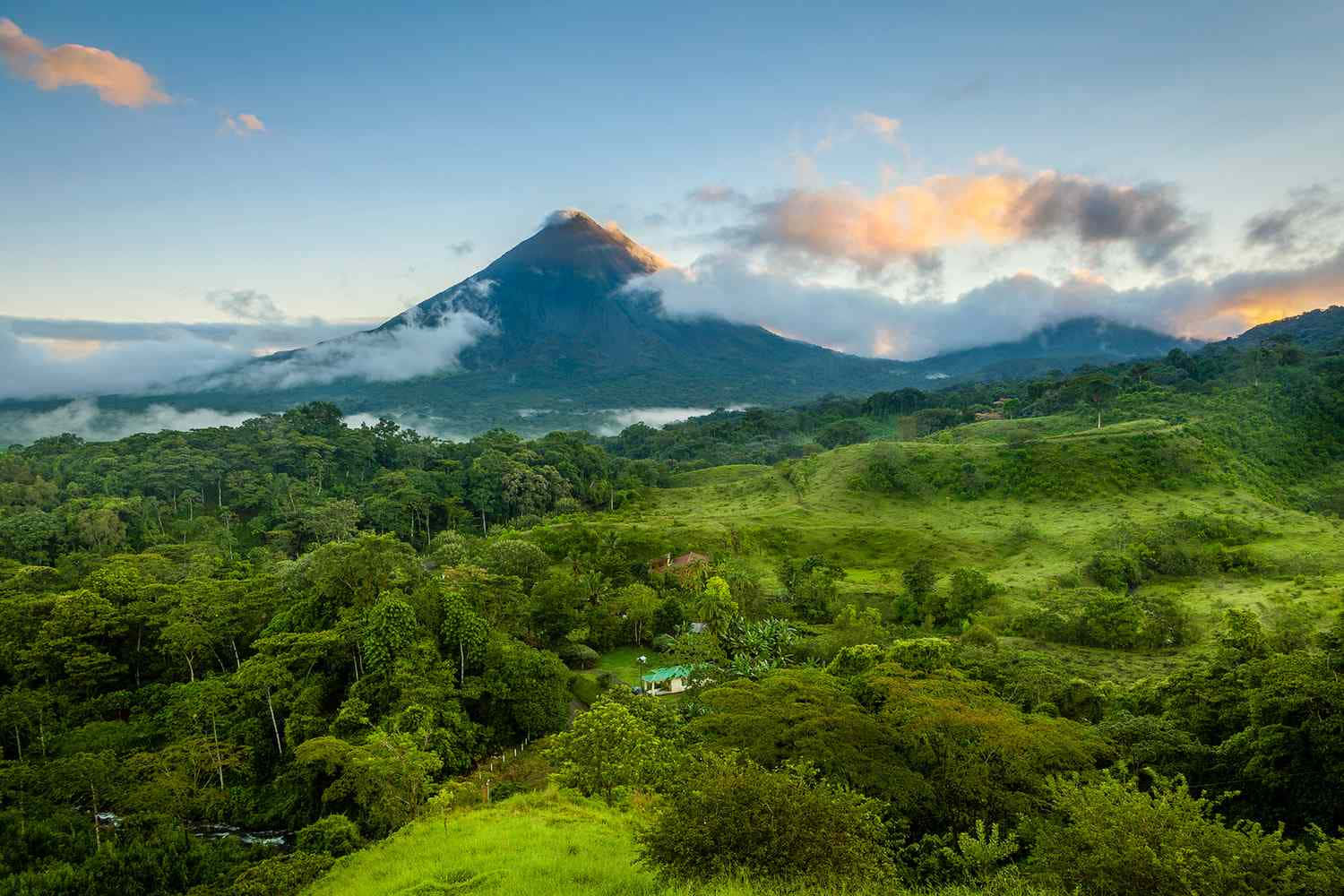 哥斯大黎加 桌布 壁紙 — 一座雄偉的火山在金色日出時升起於霧氣瀰漫的綠色山丘和茂密叢林之上。