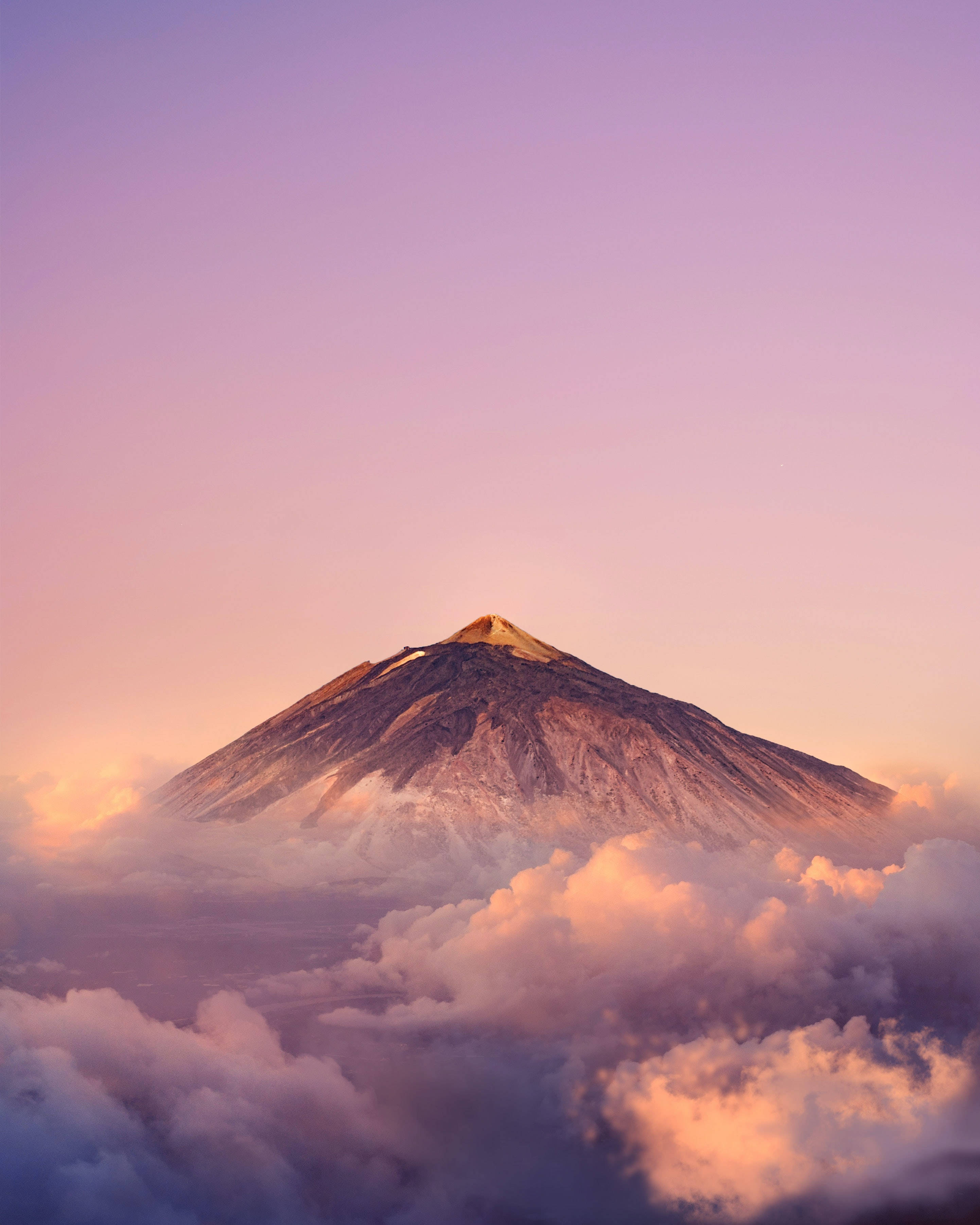 雲朵 桌布 壁紙 — 一座雄偉的火山山峰從柔軟的雲層中浮現，沐浴在柔和的粉紅色和紫色日出光中。