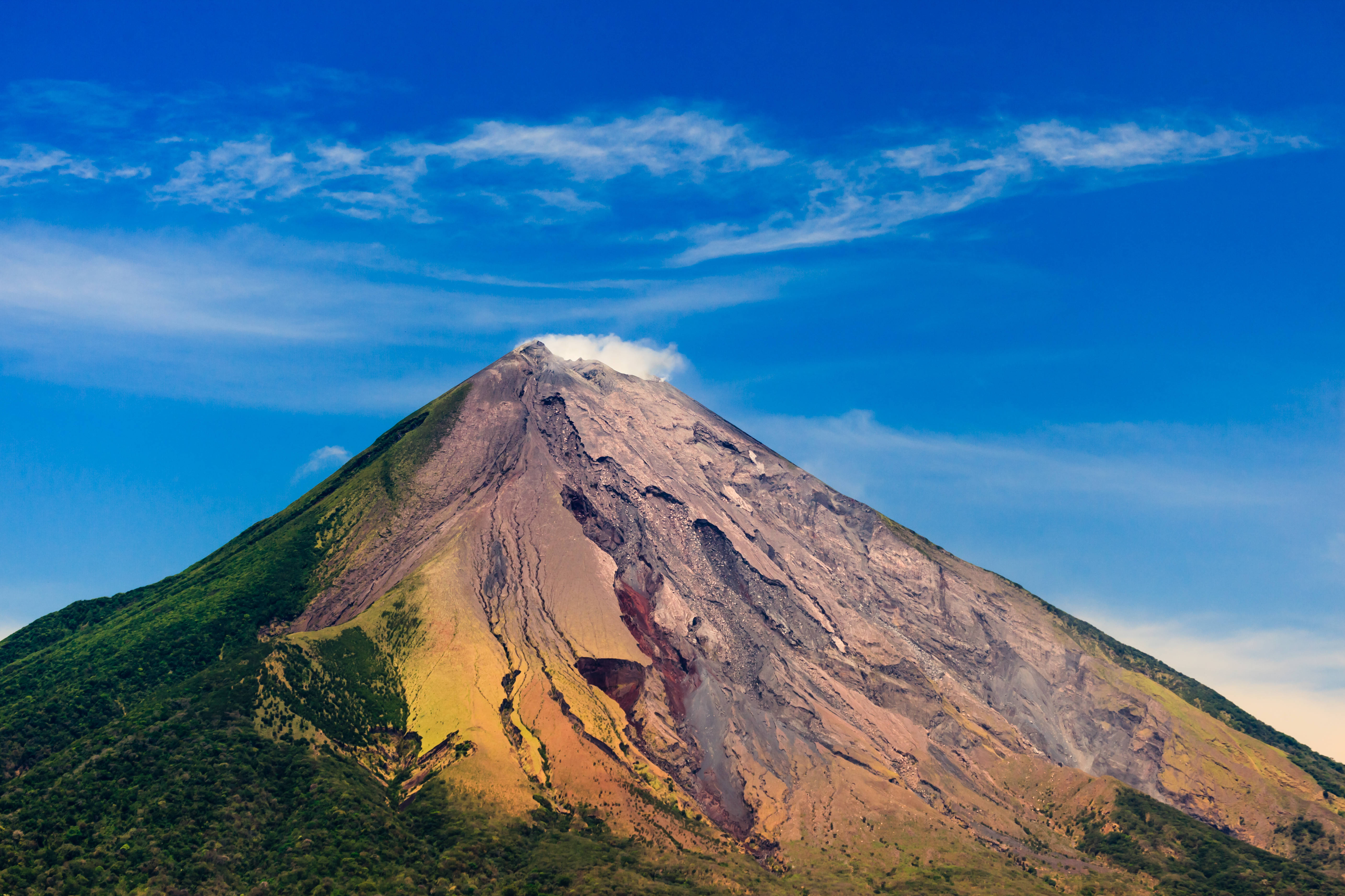Nicaragua 桌布 壁紙 — 一座雄偉的火山山脈，擁有茂密的綠色山坡和崎嶇的灰燼覆蓋的山脊，在帶有縷縷雲彩的鮮豔藍天下聳立。