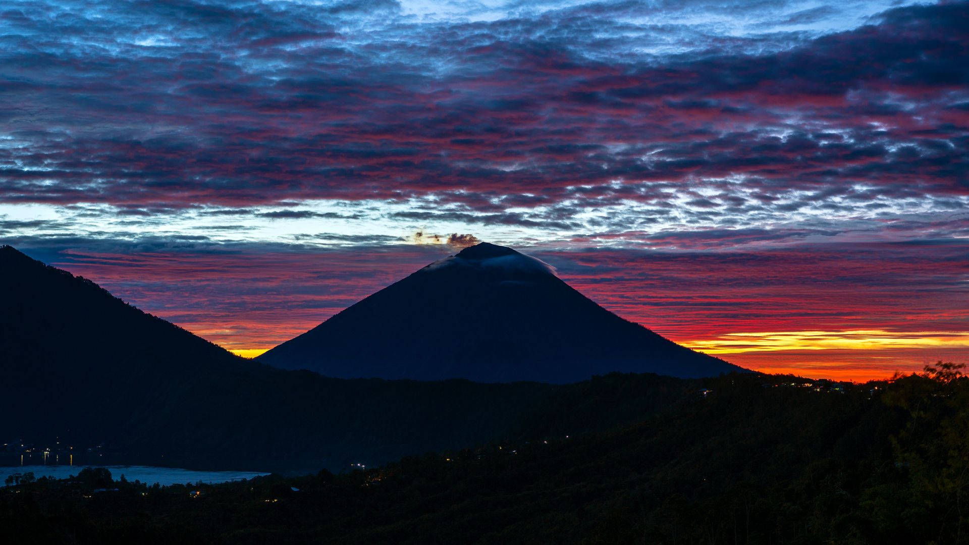 iPhone 桌布 壁紙 — 一座雄偉的火山山脈剪影矗立在火紅的日落天空前，天空佈滿紫色、橙色和藍色的雲彩。