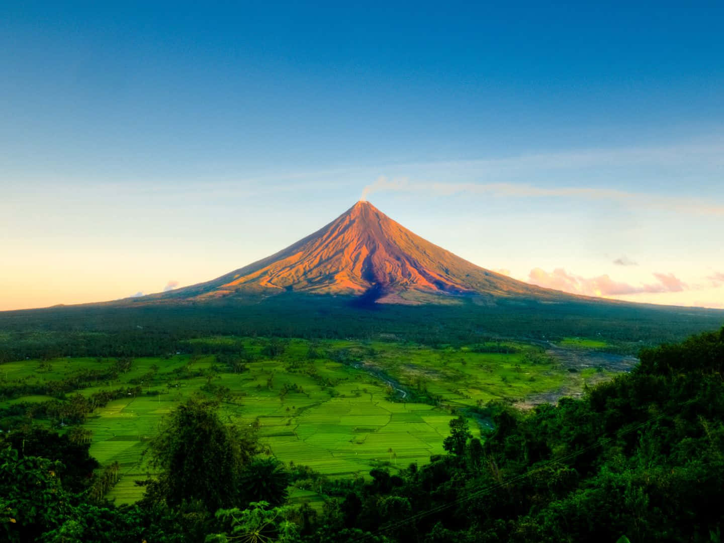 1440x1080 桌布 壁紙 — 一座雄偉的火山沐浴在金色的日出光芒中，在晴朗的藍天下，聳立在茂密的綠色稻田和茂密的森林之上。