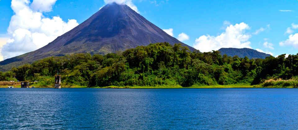 Liberia 桌布 壁紙 — 一座雄偉的火山聳立在茂密的綠色森林後面，森林毗鄰平靜的藍色湖泊，上方是明亮的藍天和蓬鬆的白雲。