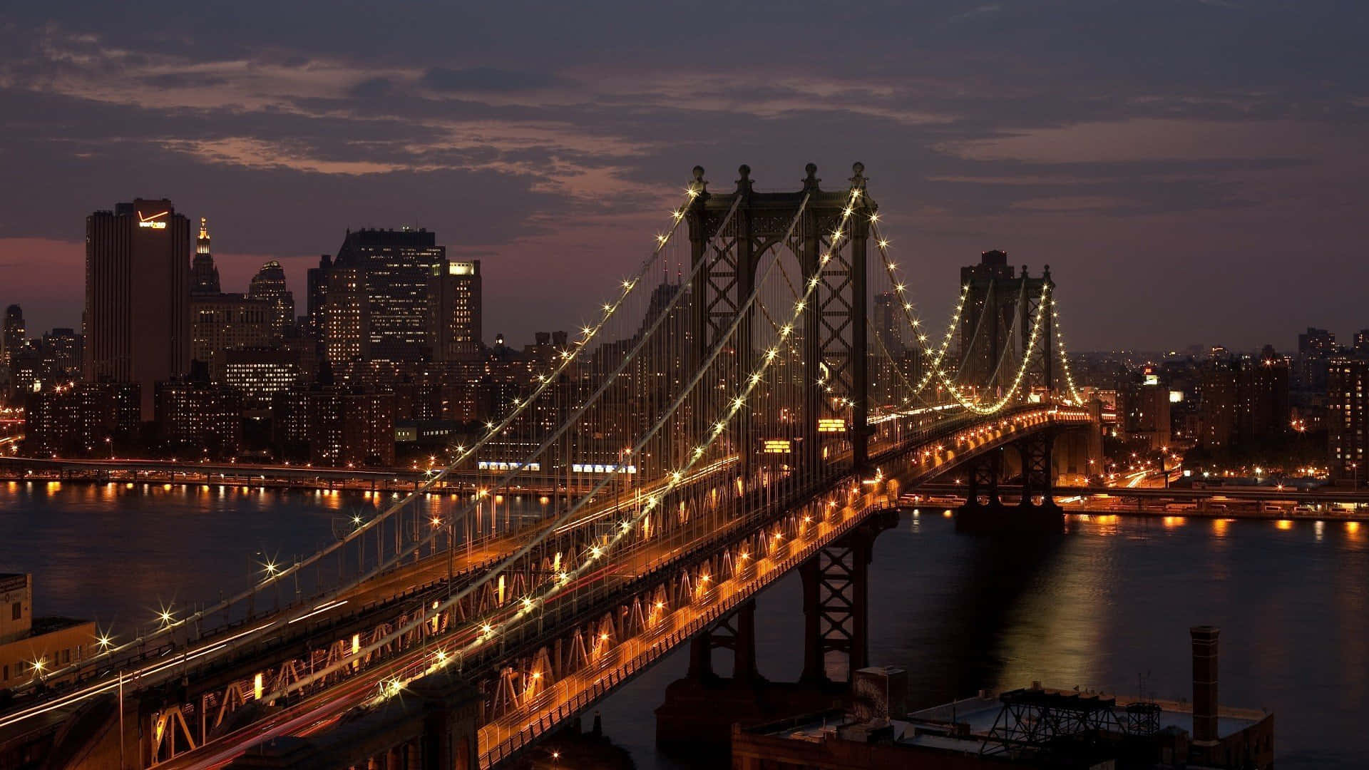 bridge 桌布 壁紙 — 一座雄偉的照明吊橋在暮色中橫跨黑暗的河流，背後是閃爍的城市天際線。