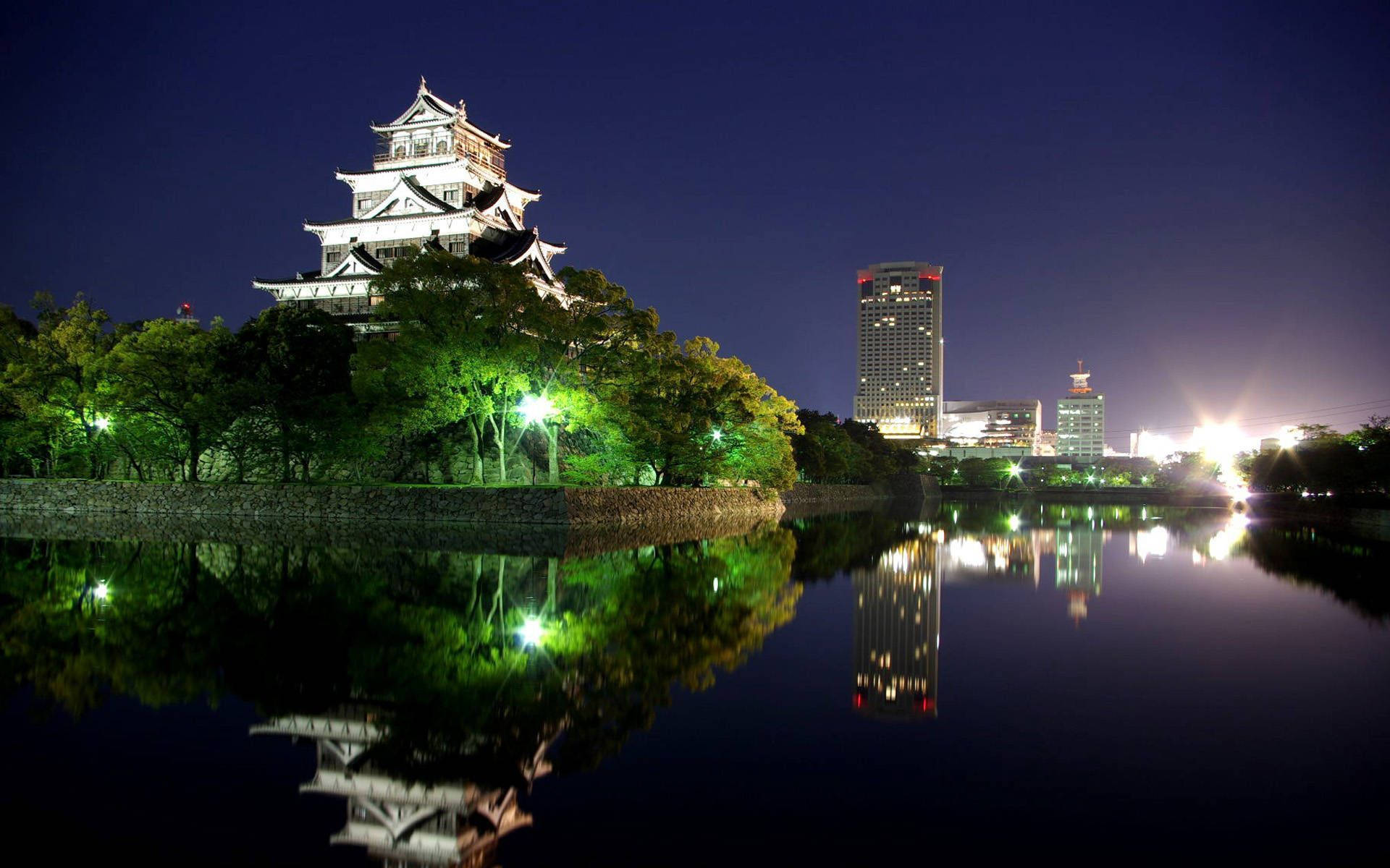 Osaka Castle 桌布 壁紙 — 一座雄偉的燈光城堡在平靜的水面上倒映，旁邊是閃爍的現代摩天大樓，在深邃的夜空下。