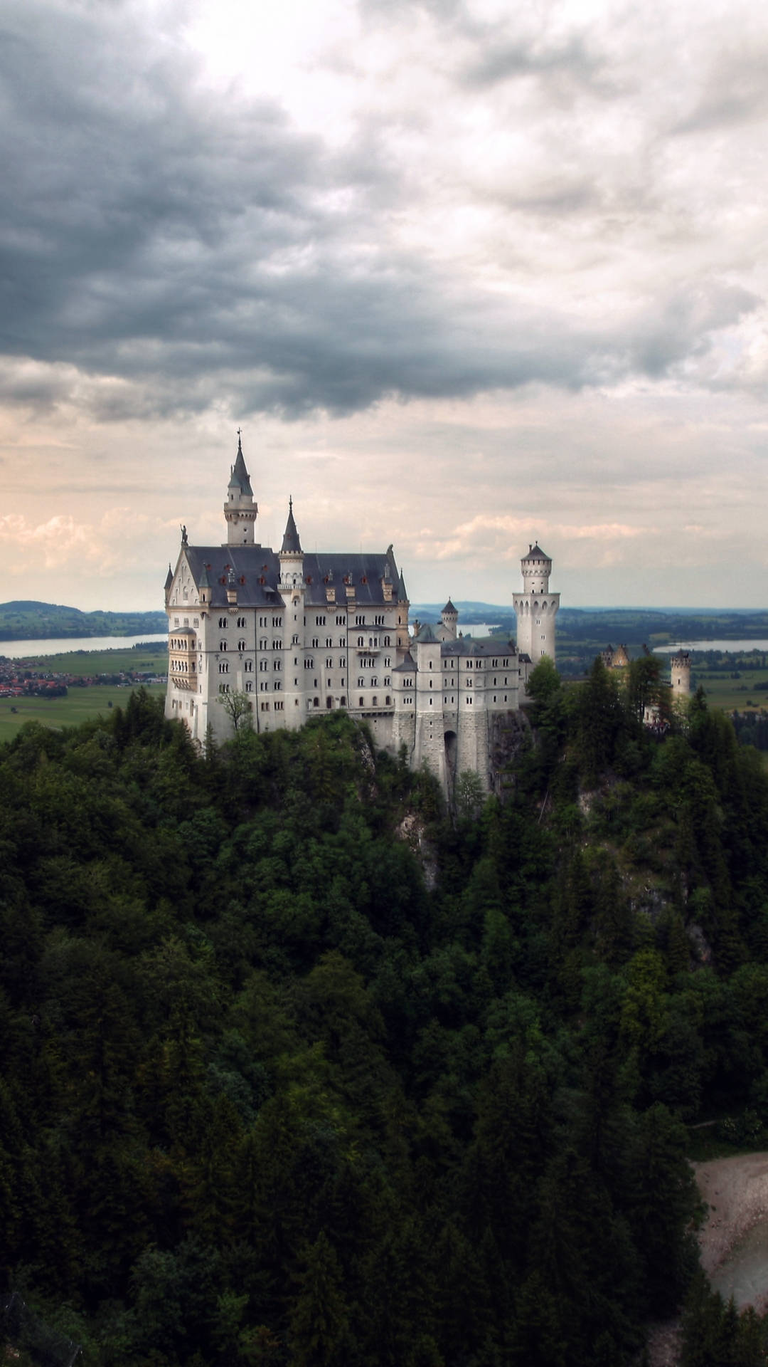 Neuschwanstein Castle 桌布 壁紙 — 一座雄偉的白色城堡，尖塔聳立在鬱鬱蔥蔥的綠色山丘上，天空戲劇性地多雲。
