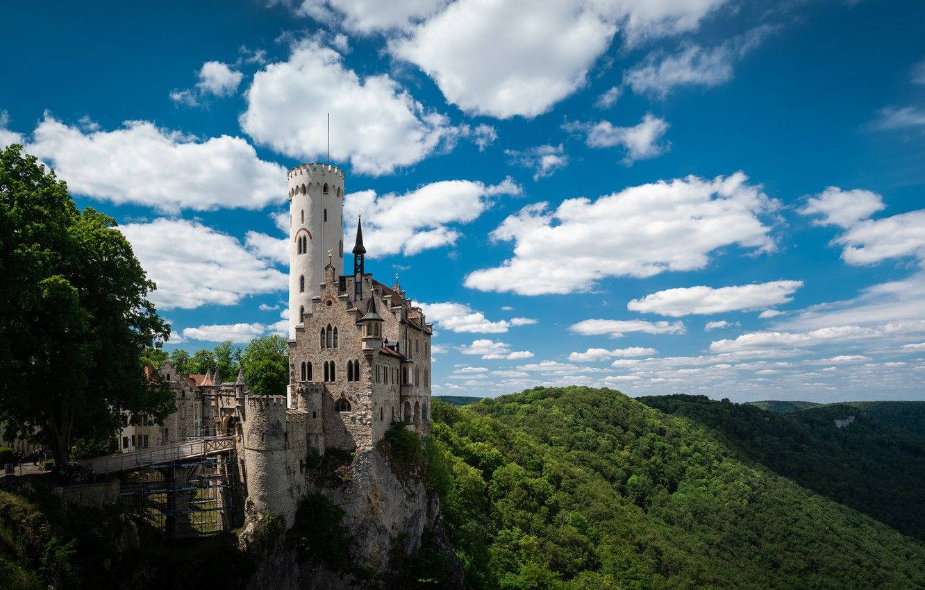 Liechtenstein 桌布 壁紙 — 一座雄偉的白色城堡坐落在懸崖上，俯瞰著鬱鬱蔥蔥的綠色山谷，上方是明亮的藍天和蓬鬆的白雲。