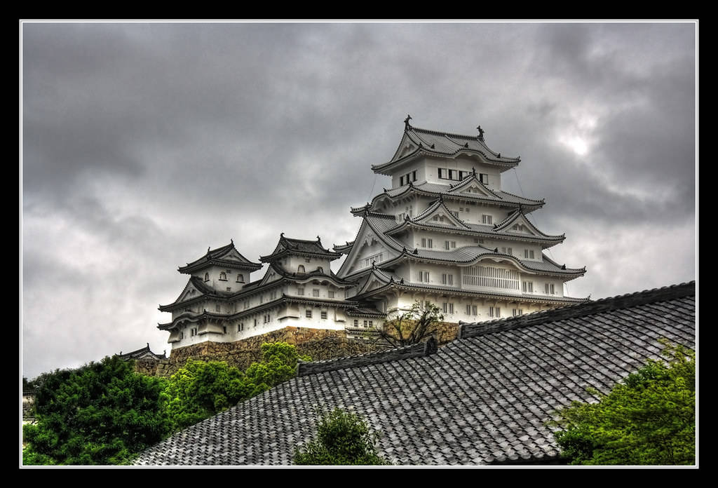 Himeji Castle 桌布 壁紙 — 一座雄偉的白色日本城堡，層疊的屋頂在戲劇性的多雲天空下，聳立在深色瓦片屋頂之上。