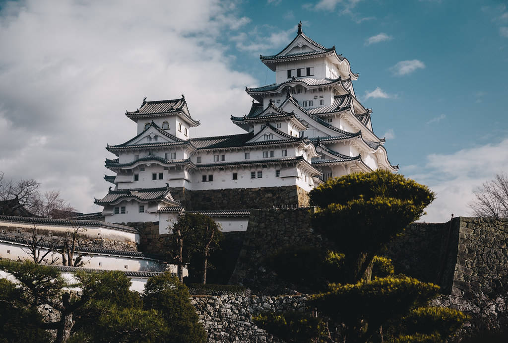 Himeji Castle 桌布 壁紙 — 一座雄偉的白色日本城堡，擁有分層屋頂，矗立在石牆上，天空部分多雲，呈現藍色。