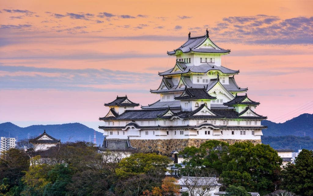Himeji Castle 桌布 壁紙 — 一座雄偉的白色日本城堡，擁有層疊屋頂，矗立在山丘上，夕陽西下，周圍環繞樹木和遠山，天空呈現粉紅和橙色。