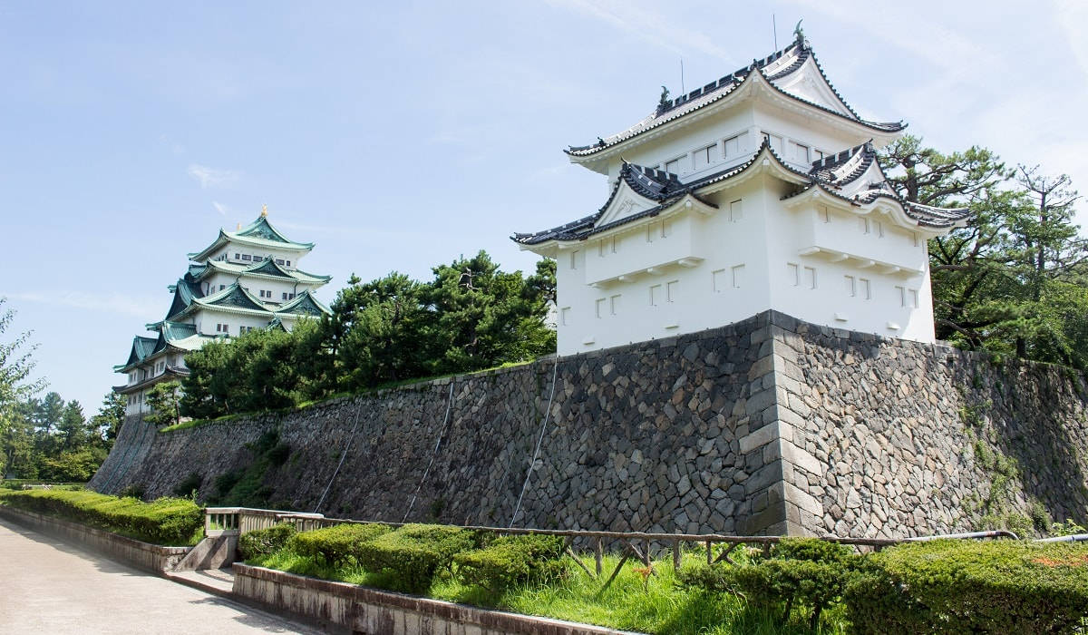 Nagoya 桌布 壁紙 — 一座雄偉的白色日本城堡，擁有綠色屋頂，矗立在石牆上，周圍環繞著茂密的綠樹，天空湛藍。