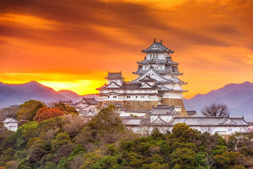Himeji Castle 桌布 壁紙 — 一座雄偉的白色日本城堡坐落在山頂上，沐浴在熾熱的橙色日落光芒中，背景是群山。