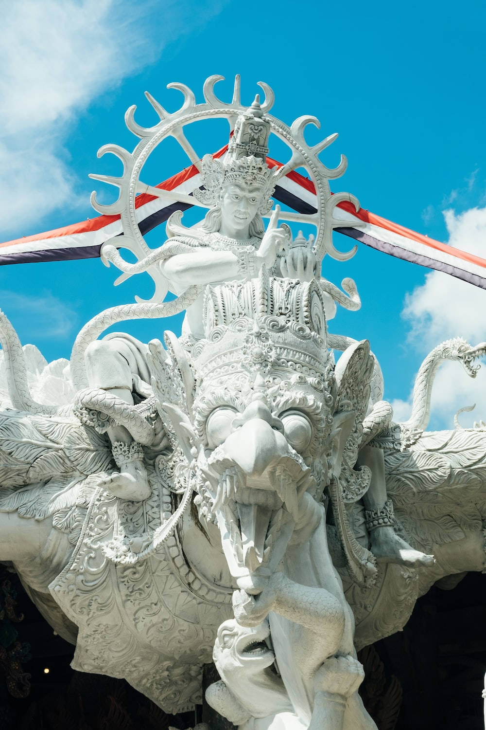 lord shiva mobile 桌布 壁紙 — 一座雄偉的白色石雕神像，雕刻精緻，頭戴光芒四射的皇冠，在鮮豔的藍天下，襯著紅、白、藍三色旗幟。
