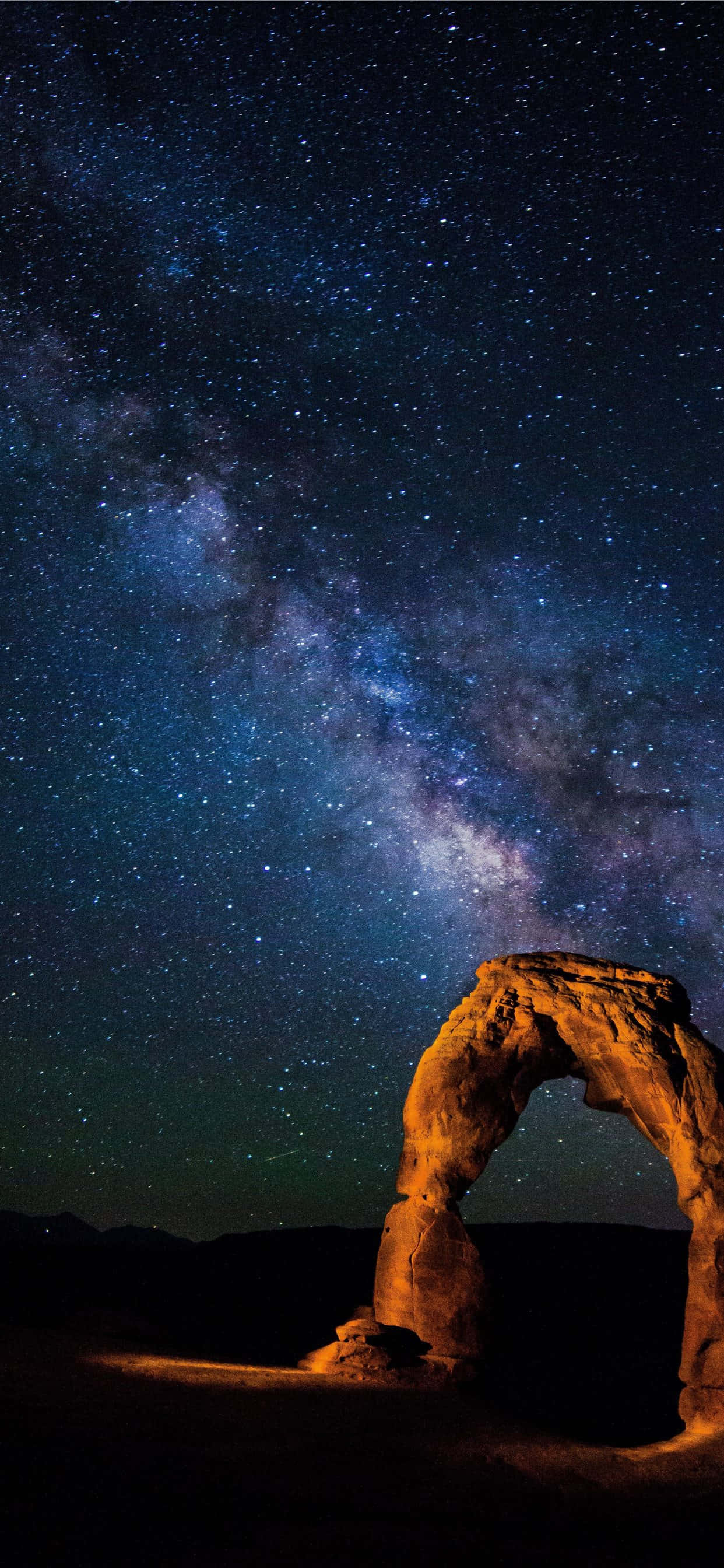夜空 桌布 壁紙 — 一座雄偉的石拱門在令人嘆為觀止的星空下溫暖地發光，星空充滿了銀河系的宇宙塵埃。