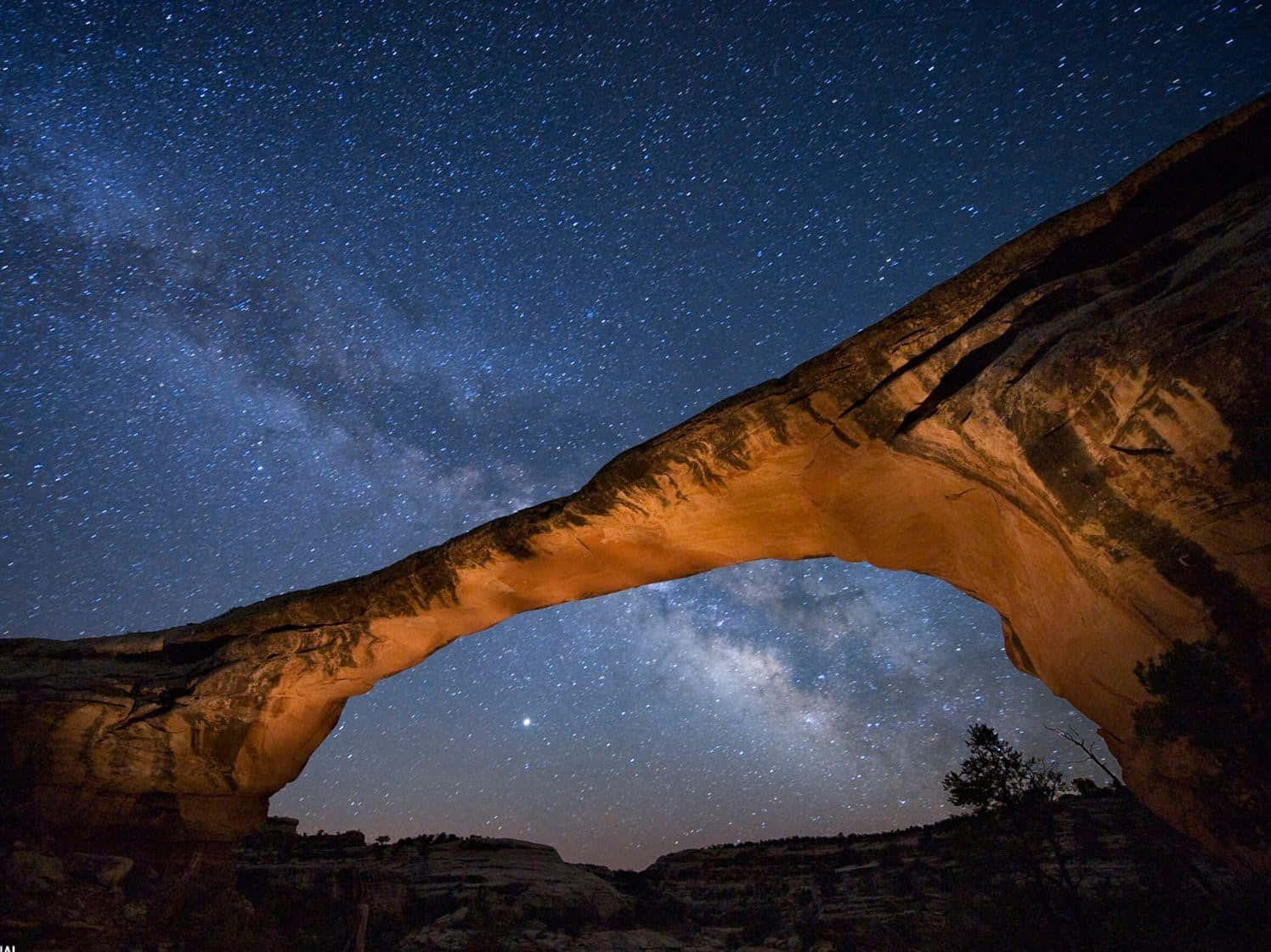 National Geographic Desktop 桌布 壁紙 — 一座雄偉的石拱門在壯麗的星空下溫暖地發光，星空充滿了銀河。