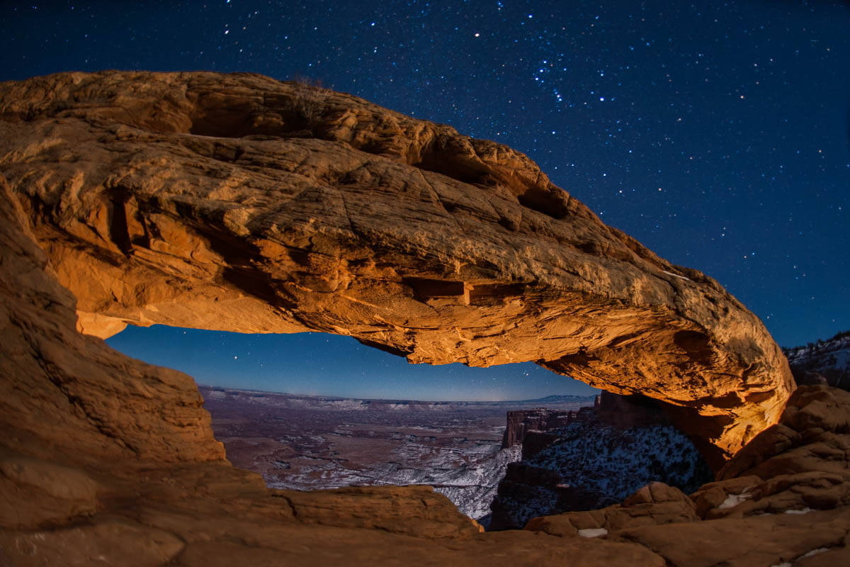 Canyonlands National Park 桌布 壁紙 — 一座雄偉的石拱門在星空下溫暖地發光，框住下方積雪的峽谷。