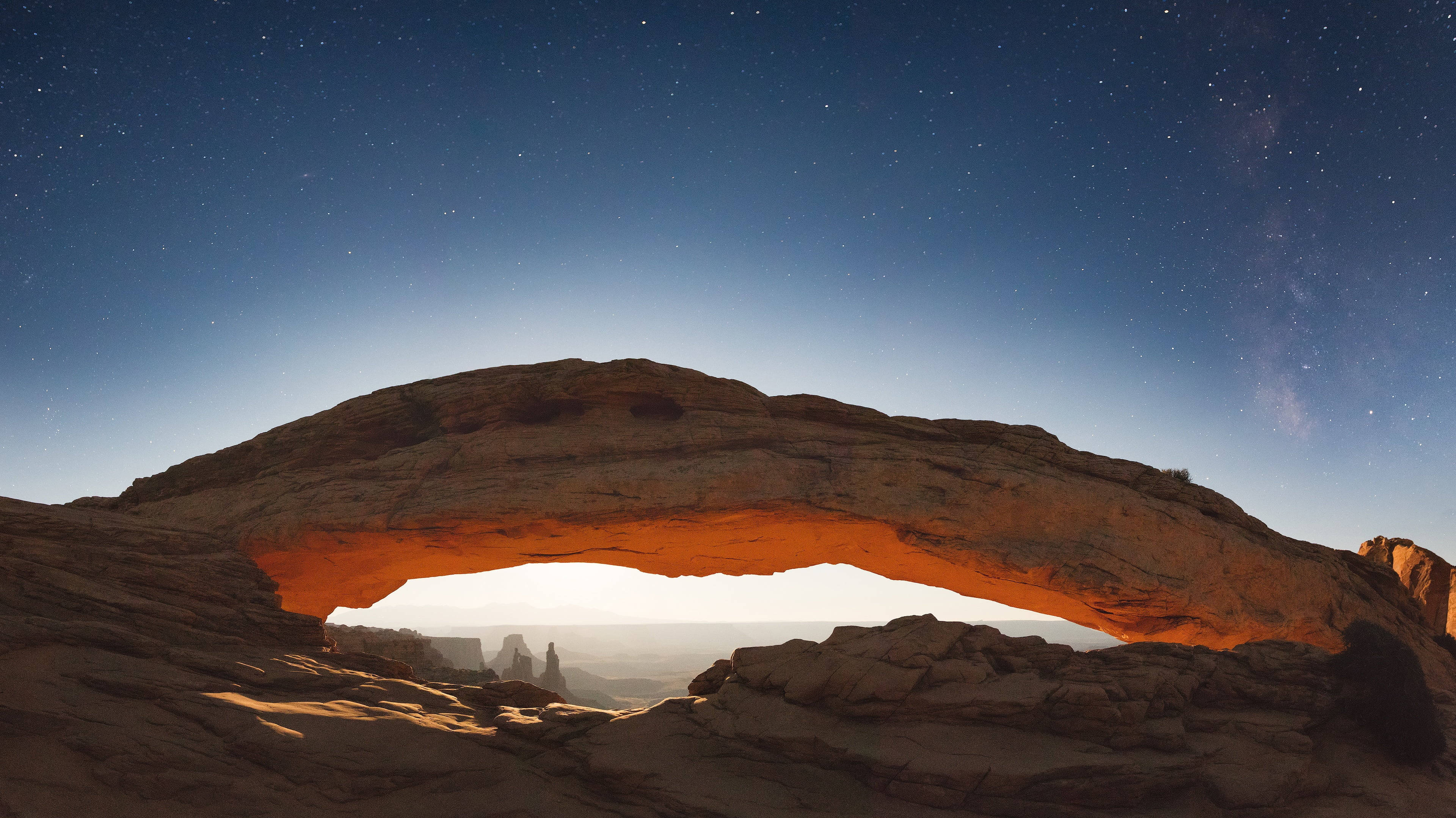 Canyonlands National Park 桌布 壁紙 — 一座雄偉的石拱門在繁星點點的夜空下溫暖地發光，銀河系清晰可見。
