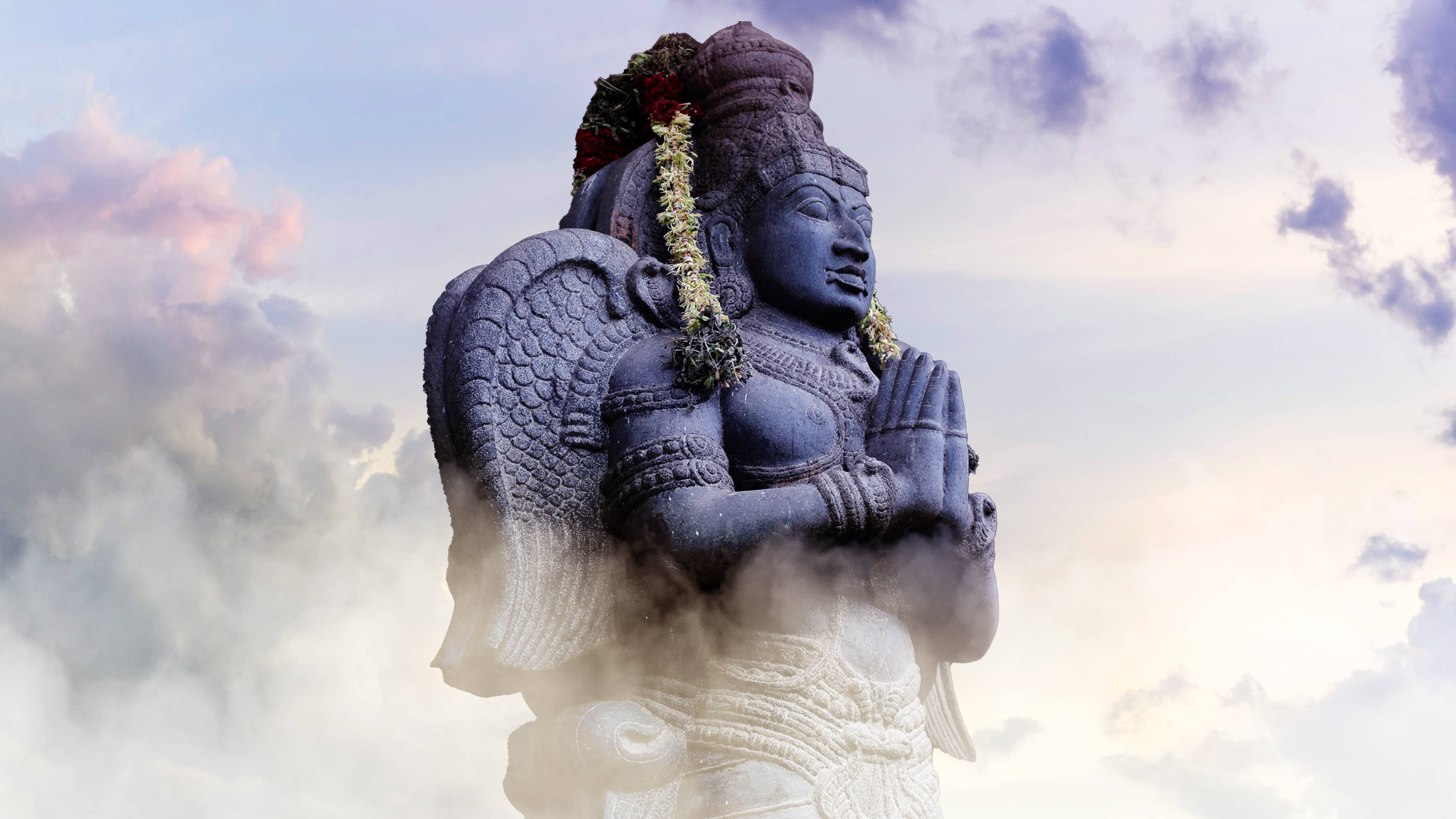 Statue Aesthetic 桌布 壁紙 — 一座雄偉的石神像，帶有翅膀和花環，在柔和的雲天背景下，底座周圍有薄霧繚繞，靜謐地矗立著。