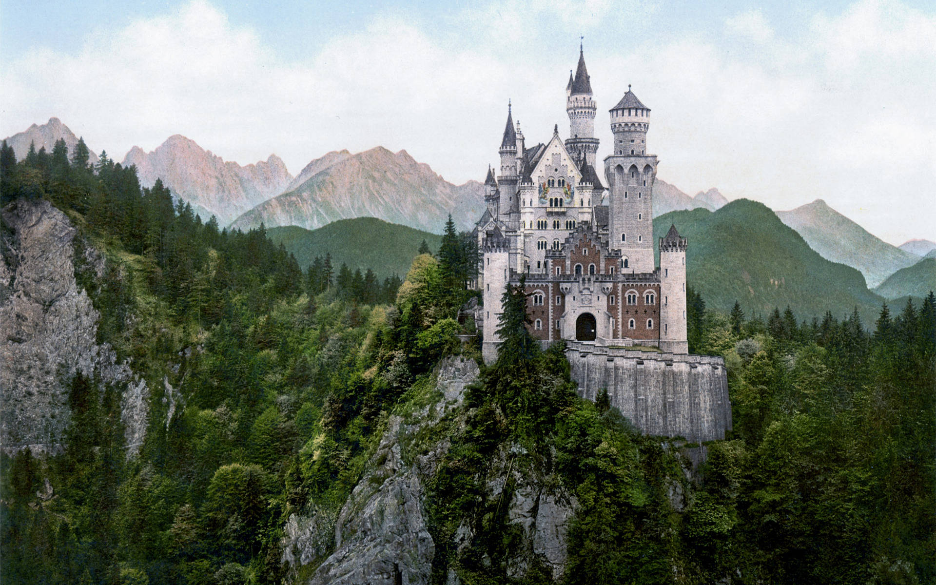 Neuschwanstein Castle 桌布 壁紙 — 一座雄偉的童話城堡坐落在岩石懸崖上，周圍環繞著茂密的綠色森林和聳立的山脈，天空柔和多雲。