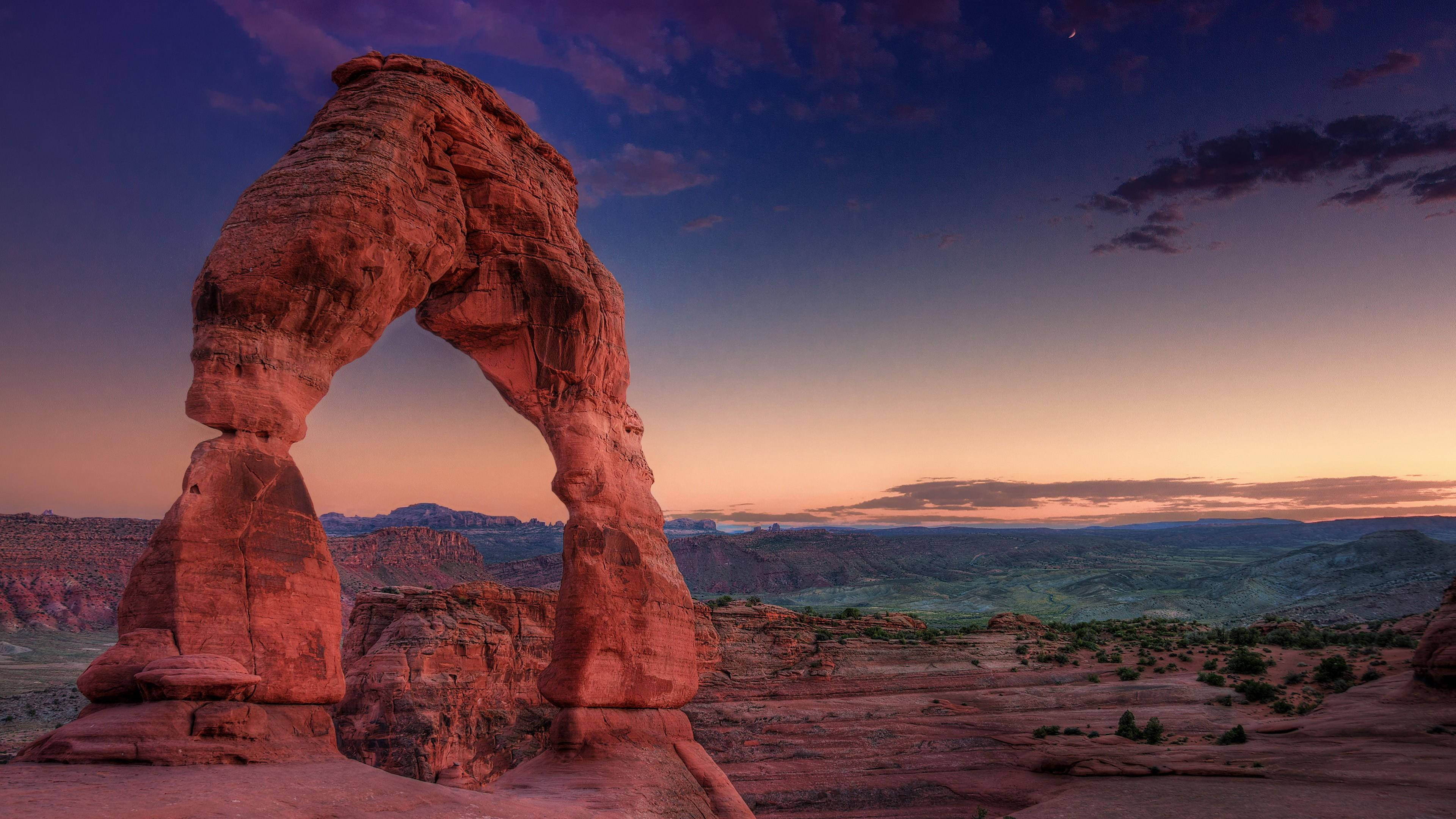 Canyonlands National Park 桌布 壁紙 — 一座雄偉的紅色砂岩拱門在溫暖的暮色色調中發光，映襯著漸變的天空、遠山和稀疏的沙漠植被。