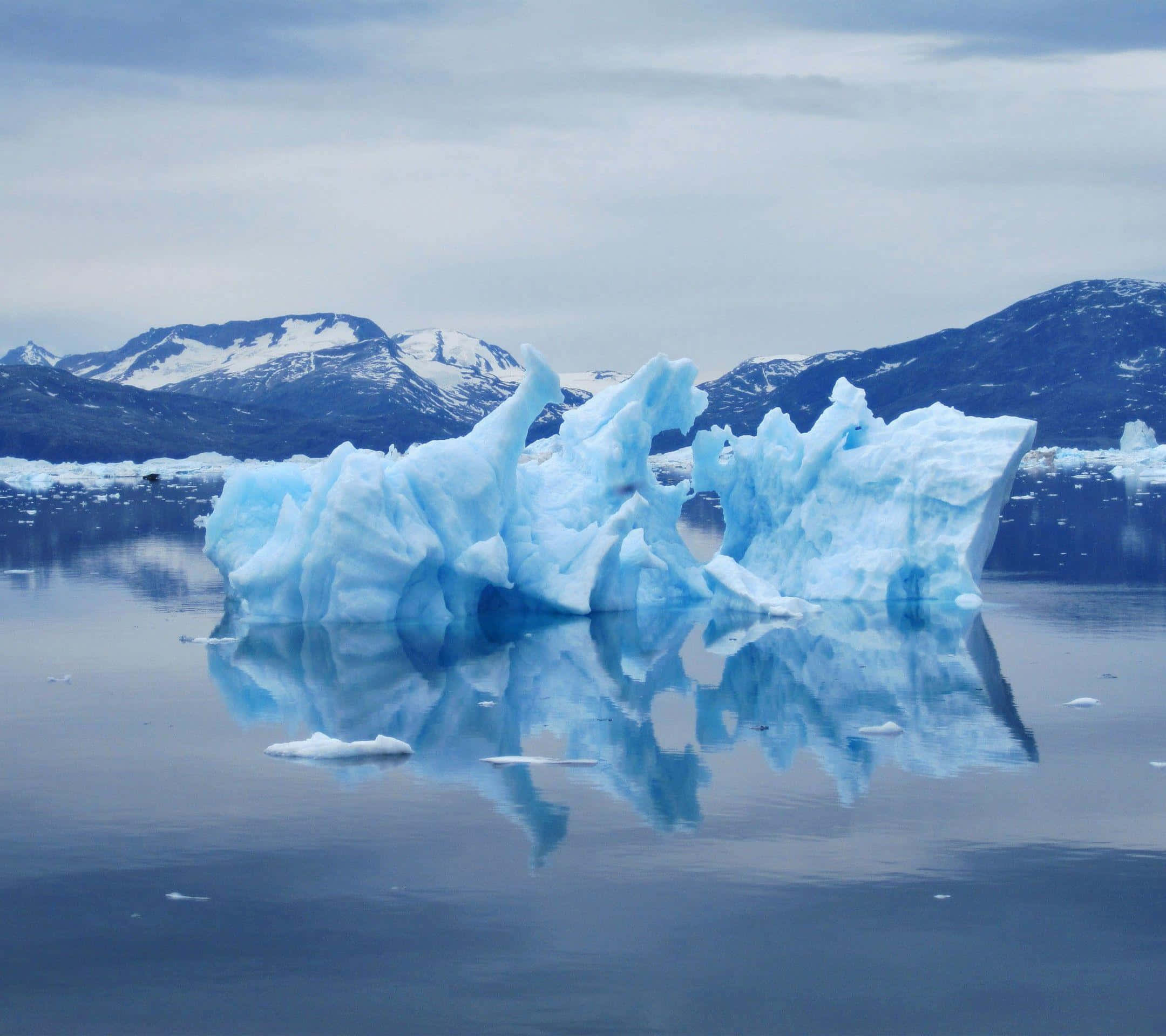 Greenland 桌布 壁紙 — 一座雄偉的藍色冰山在平靜的水面上靜靜漂浮，白雪覆蓋的山脈映照在玻璃般的水面上，天空多雲。