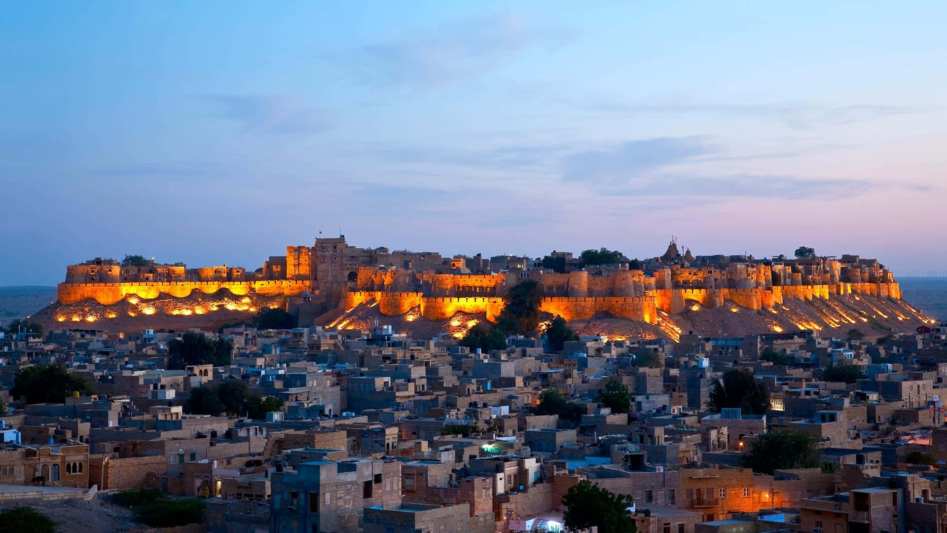 Rajasthan 桌布 壁紙 — 一座雄偉的金色堡壘矗立在山頂上，俯瞰著暮色中廣闊的古城。