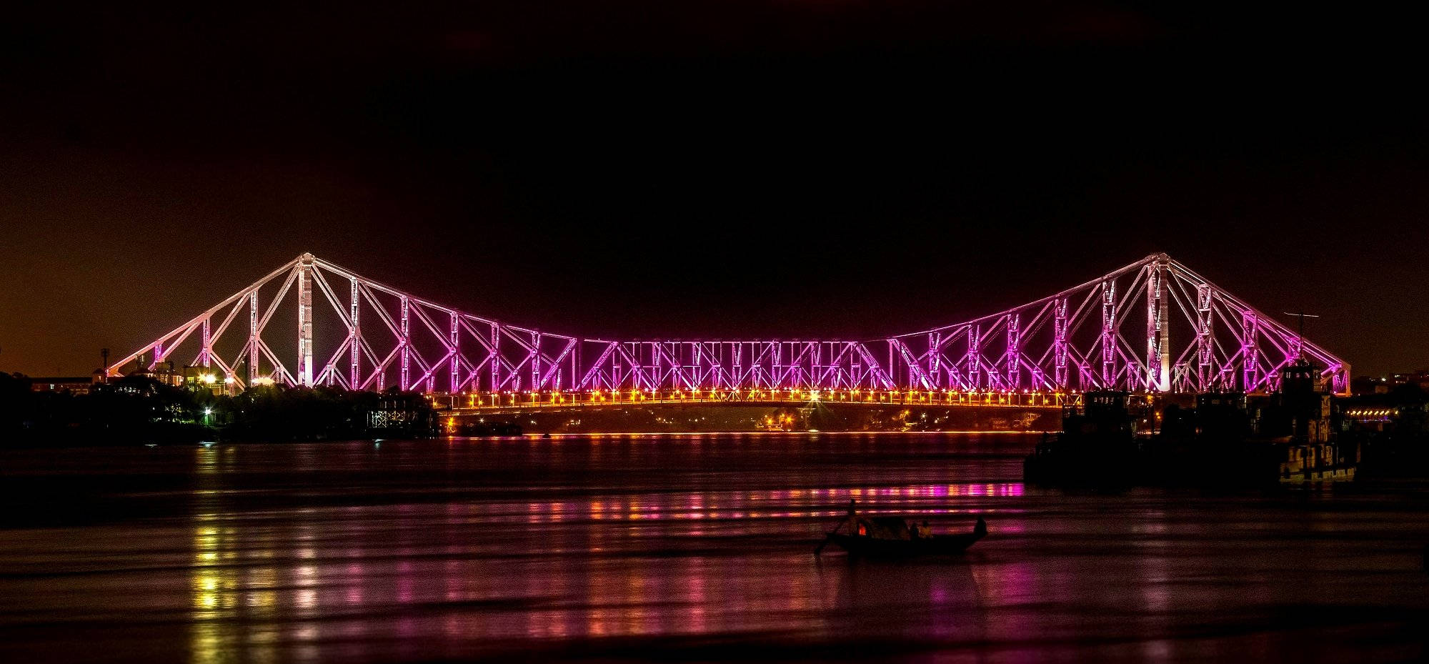 Kolkata 桌布 壁紙 — 一座雄偉的鋼桁架橋在夜間發出鮮豔的紫色光芒，照亮黑暗的河流，其倒影在水面上閃爍，一艘小船在下方漂流。